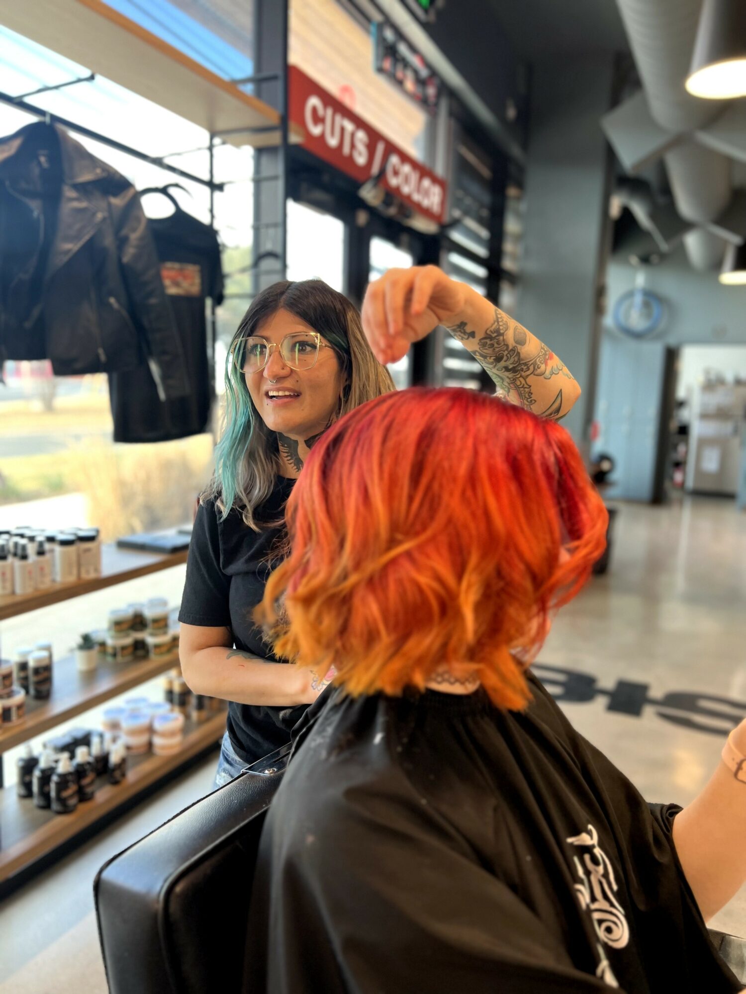 Woman with glasses and dark hair smiling, flexing her arm, in a salon with red hair and black cape, shelves behind.
