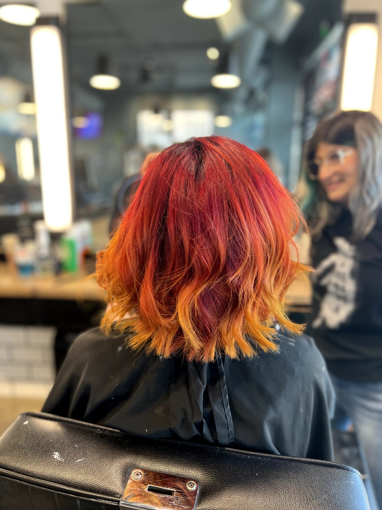 Person with vibrant red and orange wavy hair sitting in a salon chair, with a stylist smiling in the background.