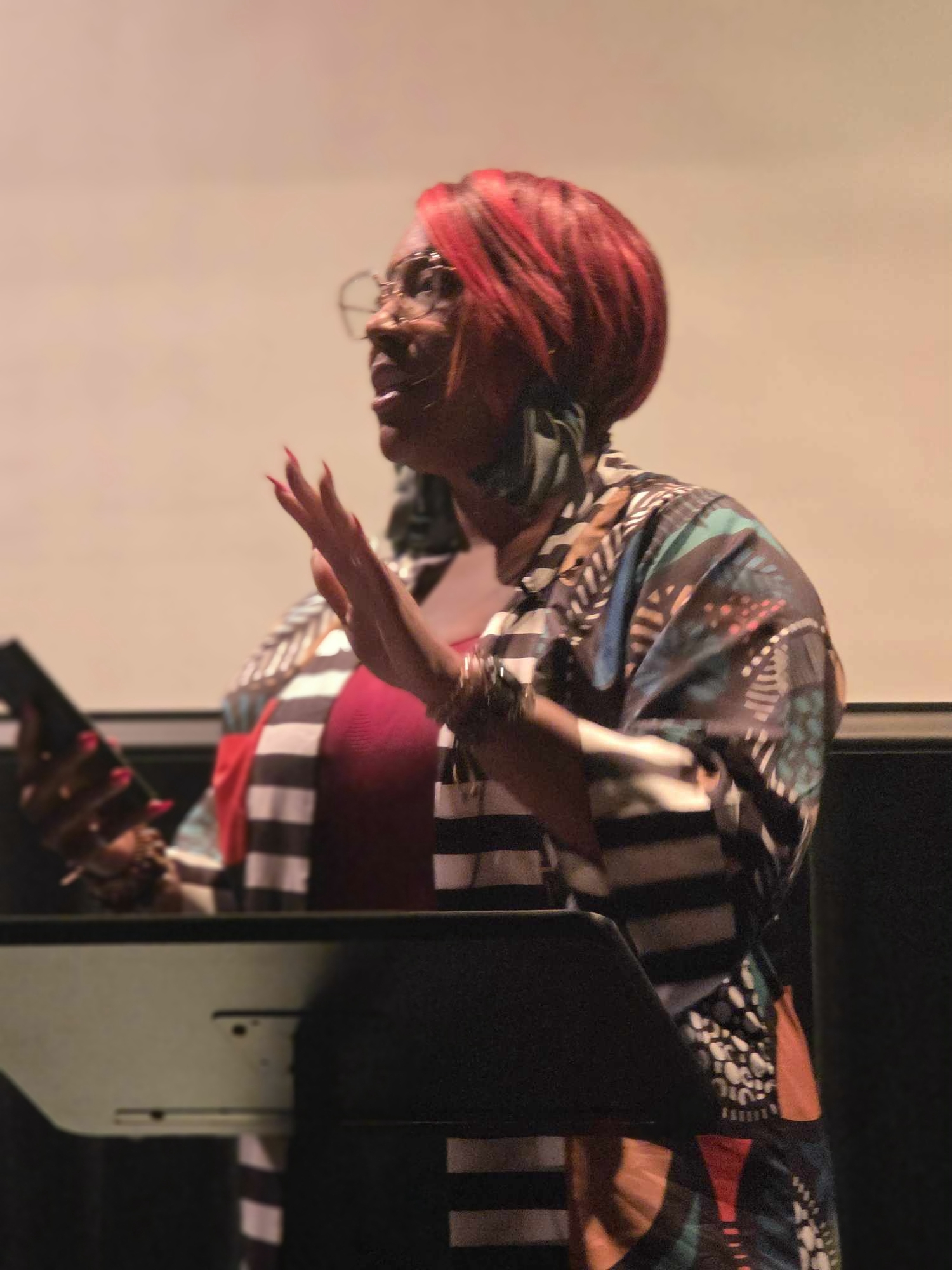 Woman with red hair, glasses, and colorful clothing, speaking or presenting, holding a device, in a dimly lit setting.