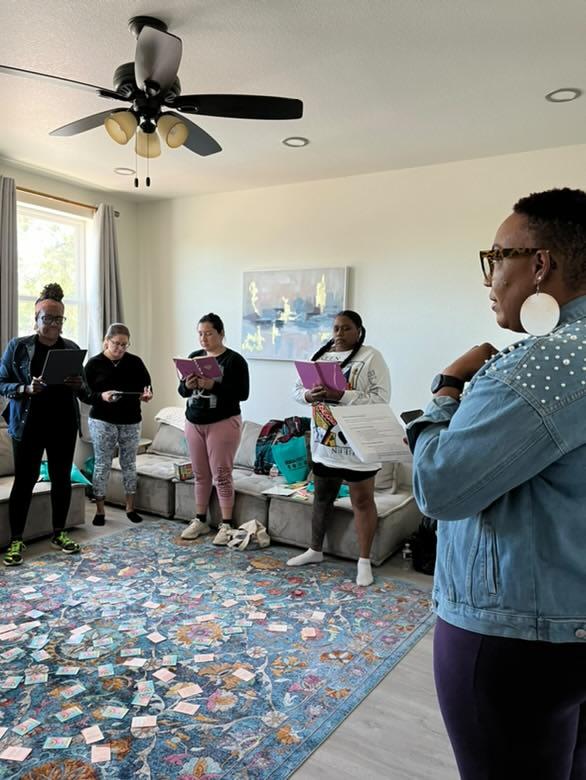 Group of five people standing in a living room, some holding notebooks, with a woman in the foreground wearing glasses and earrings.