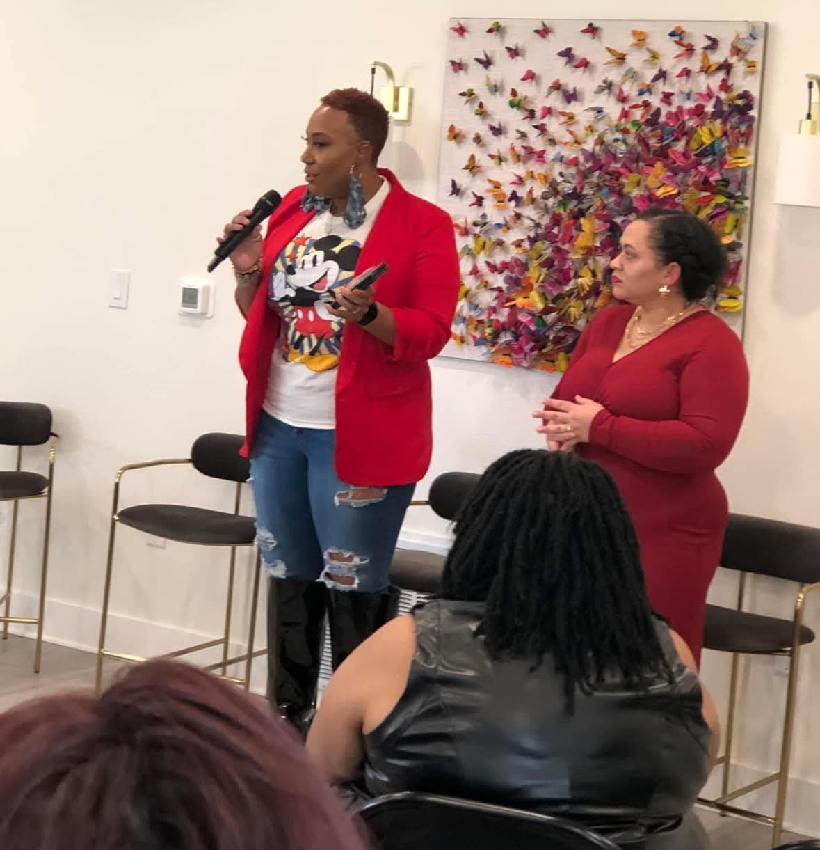 Two women speaking in front of an audience, one holding a microphone and a book, with a colorful butterfly artwork behind them.