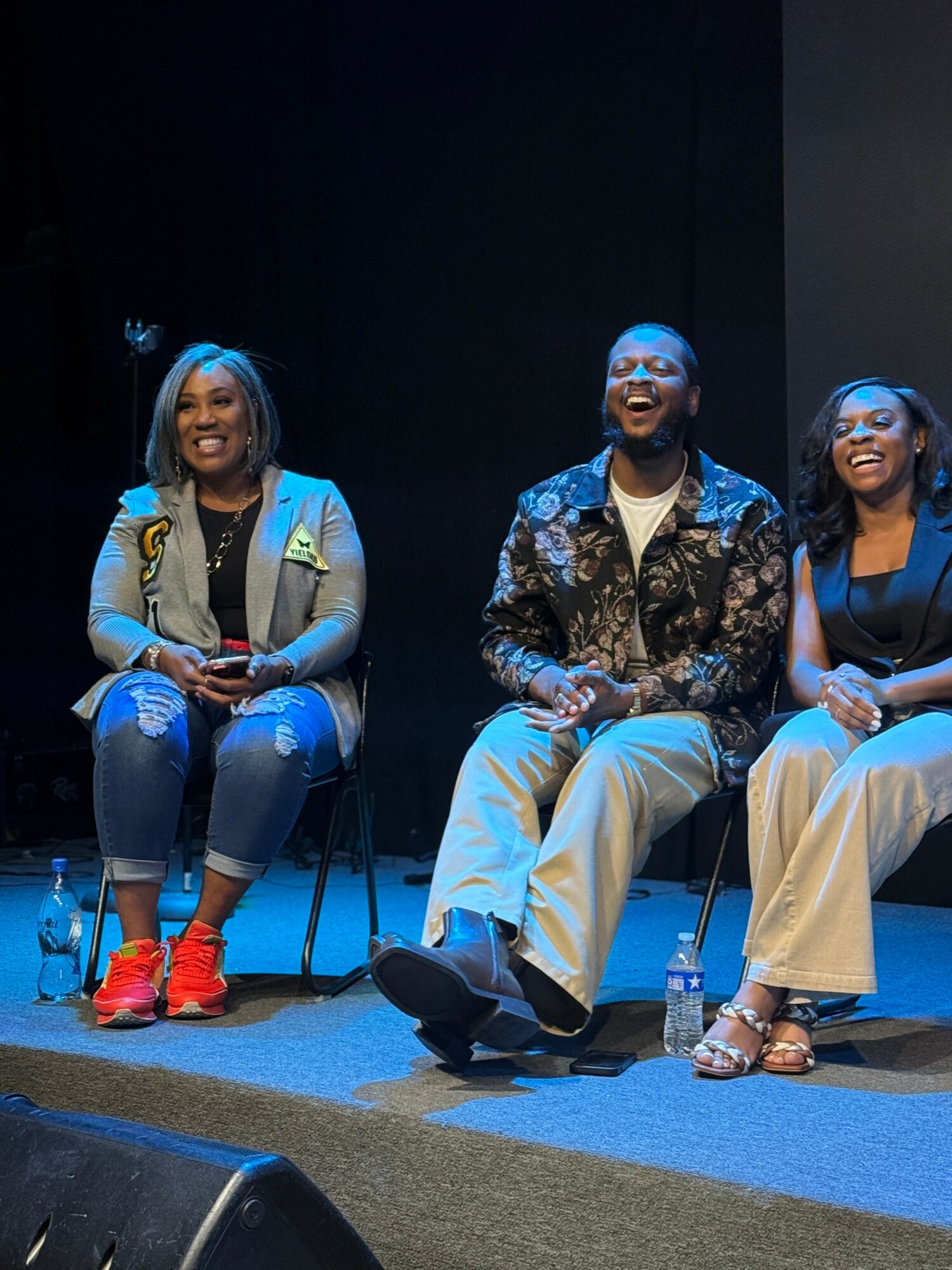 Three people sitting on chairs on stage, smiling and laughing, with water bottles on the floor, dark background.