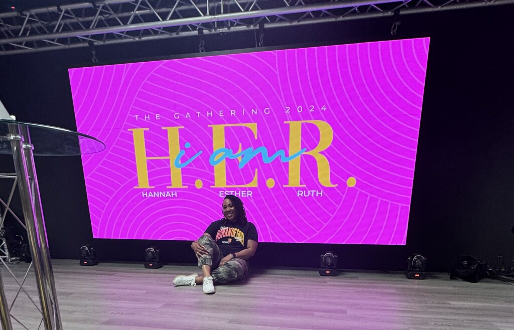 Person sitting on the floor in front of a large pink screen with yellow text, in a dark room.