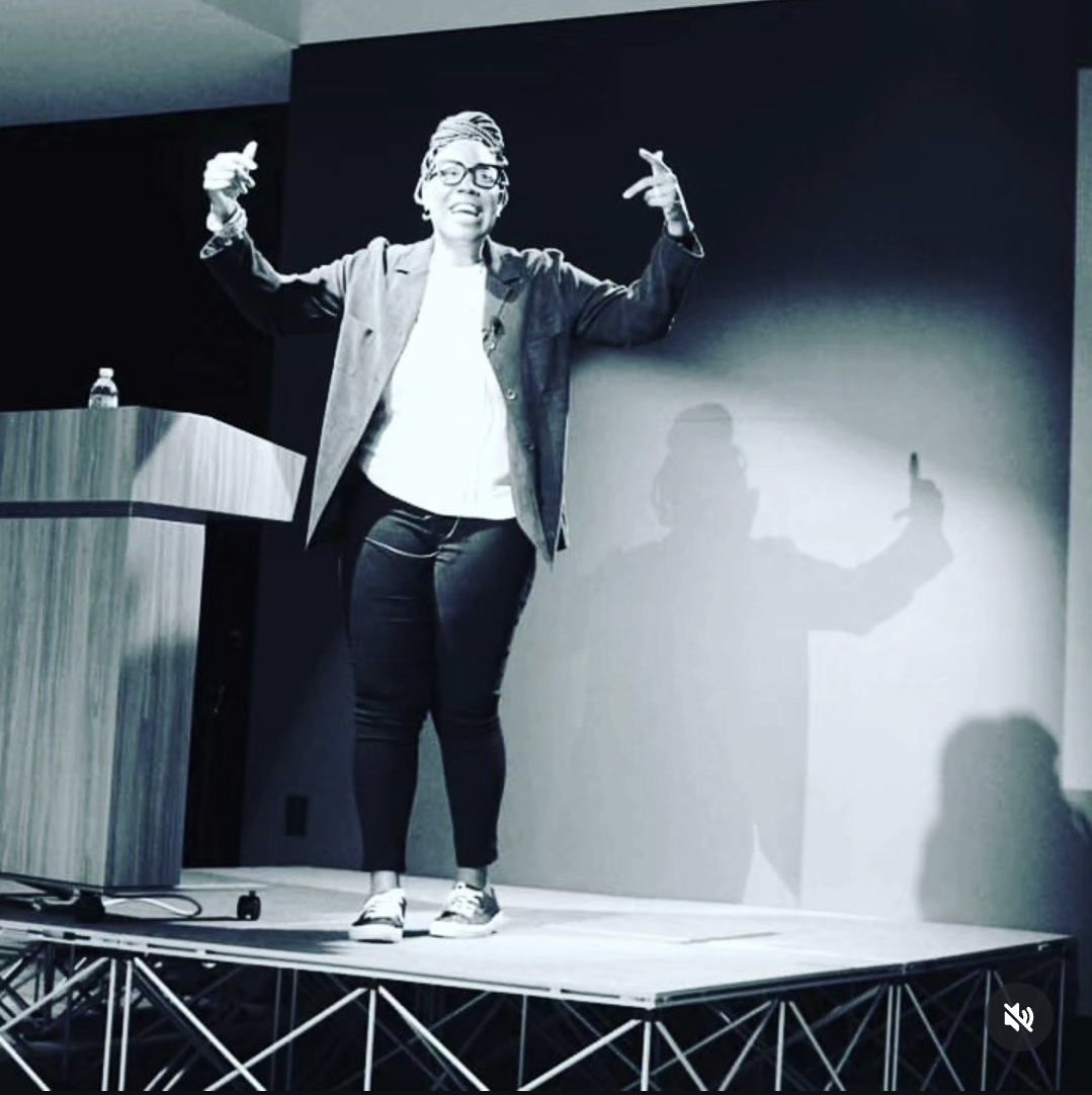 Person standing on stage with arms raised, giving a presentation or speech, shadow cast on wall behind them.