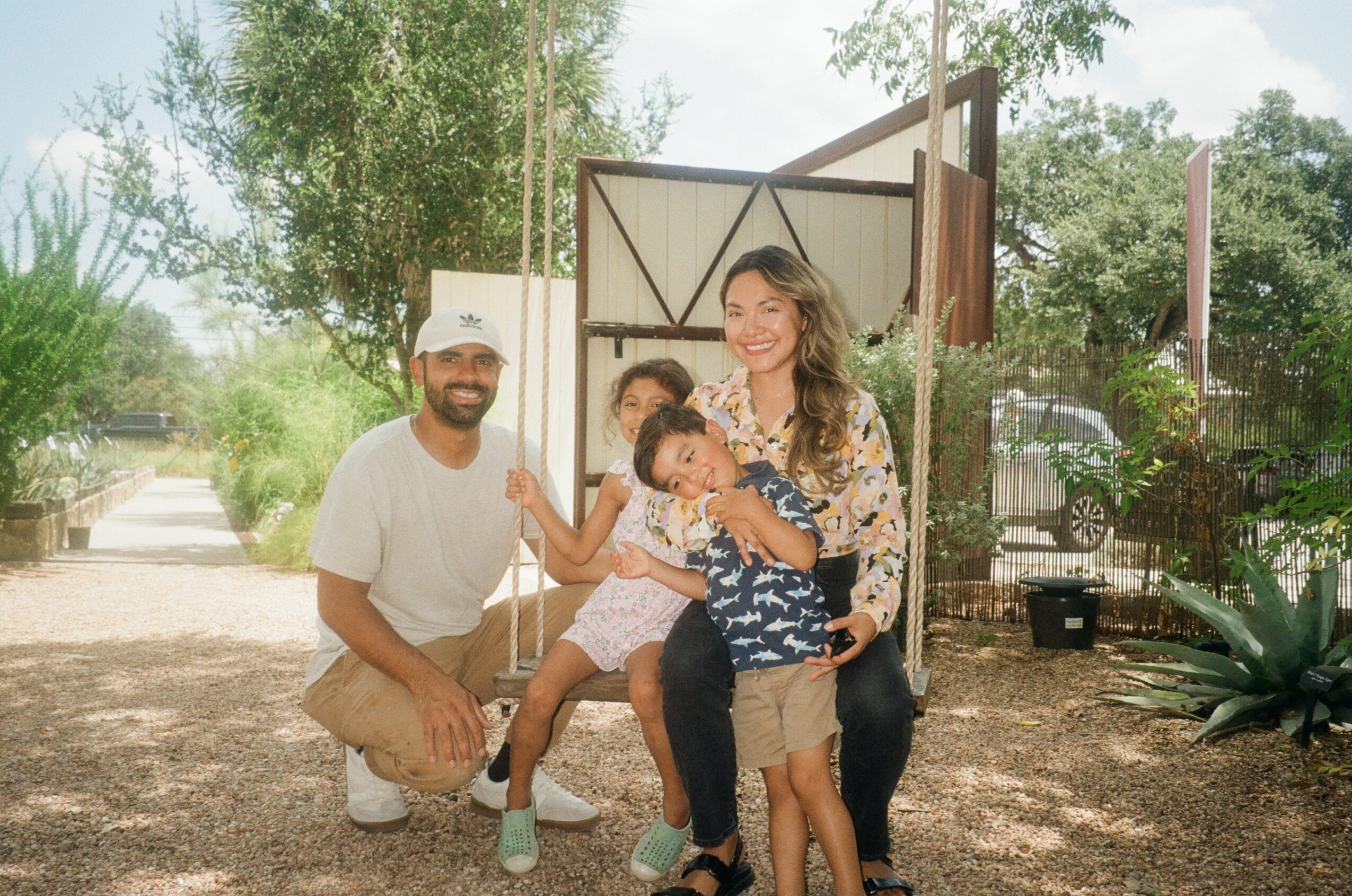 A family of four outdoors, smiling, with trees and a fence in the background.