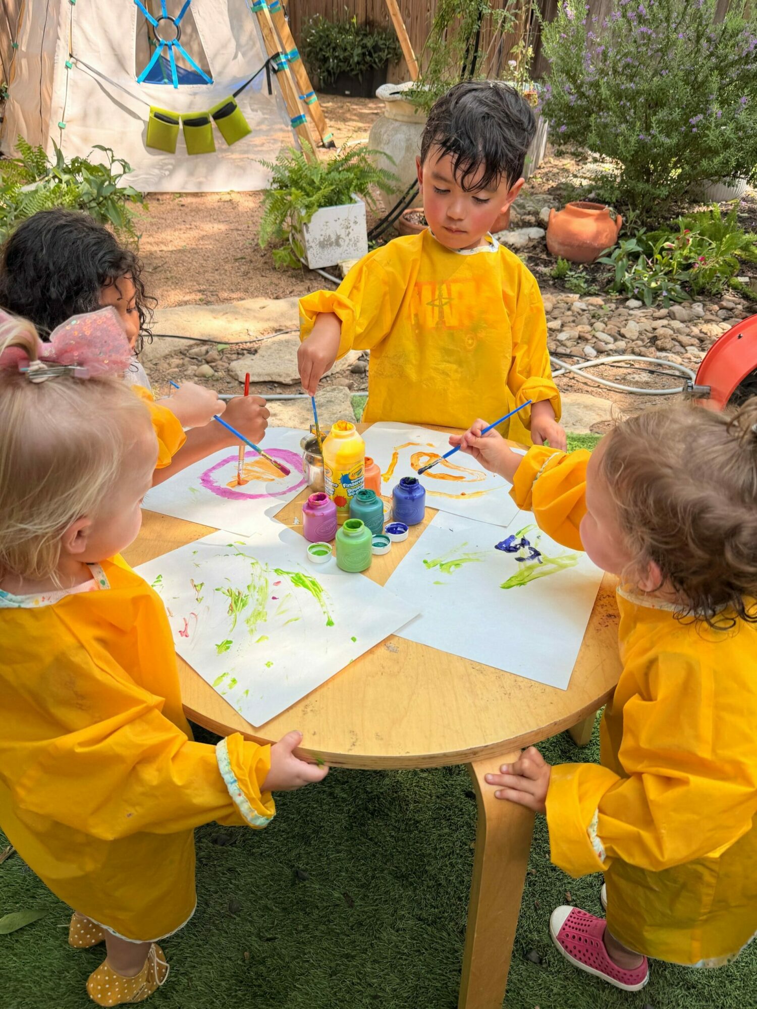 Two children in yellow smocks drawing on a large white paper outdoors, with trees and plants in the background.