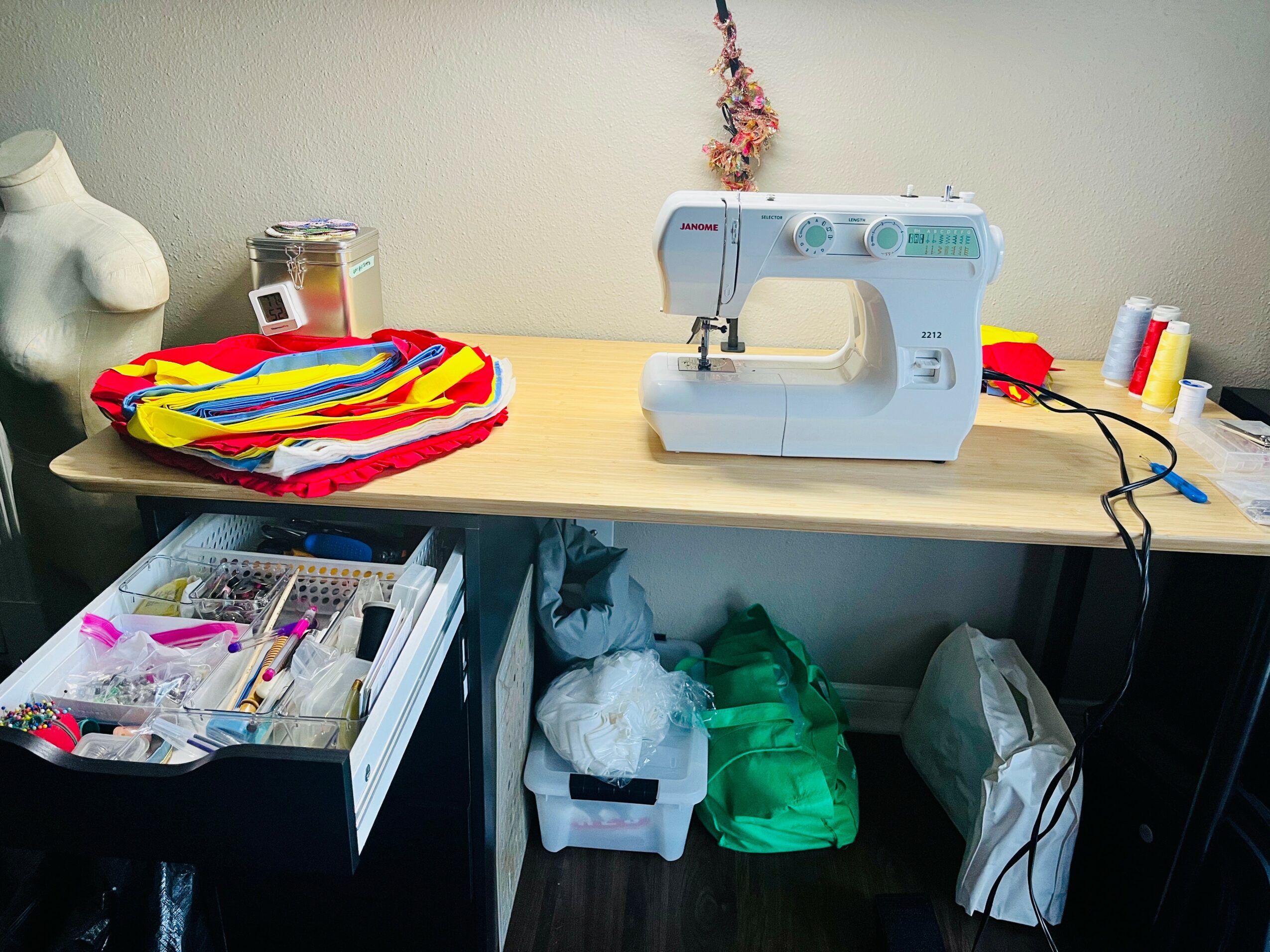 Sewing machine on a wooden table with fabric, thread, and sewing supplies, and a drawer with organized sewing tools.