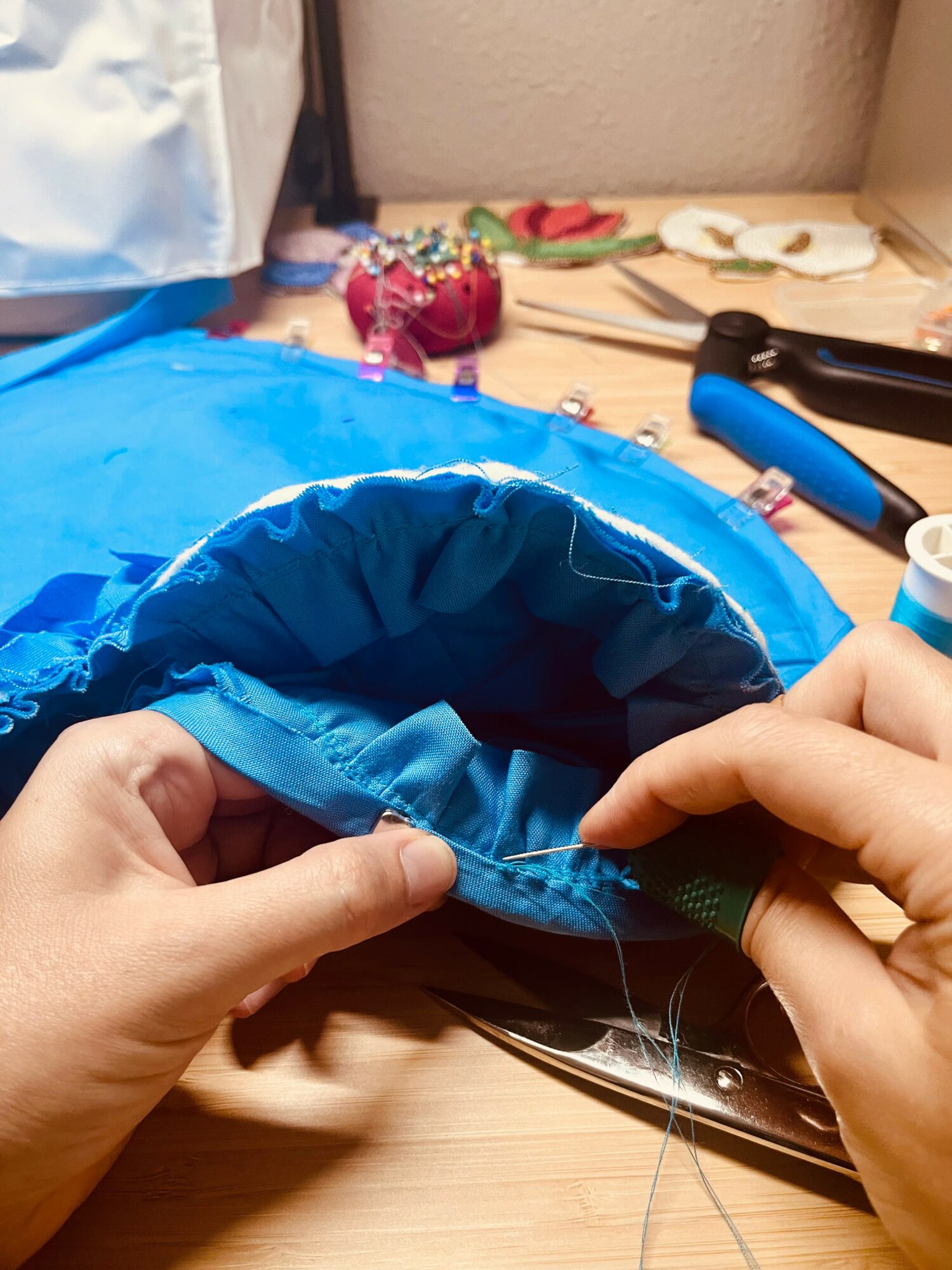Hands sewing blue fabric with a needle and thread, sewing a hem or seam, on a wooden surface.
