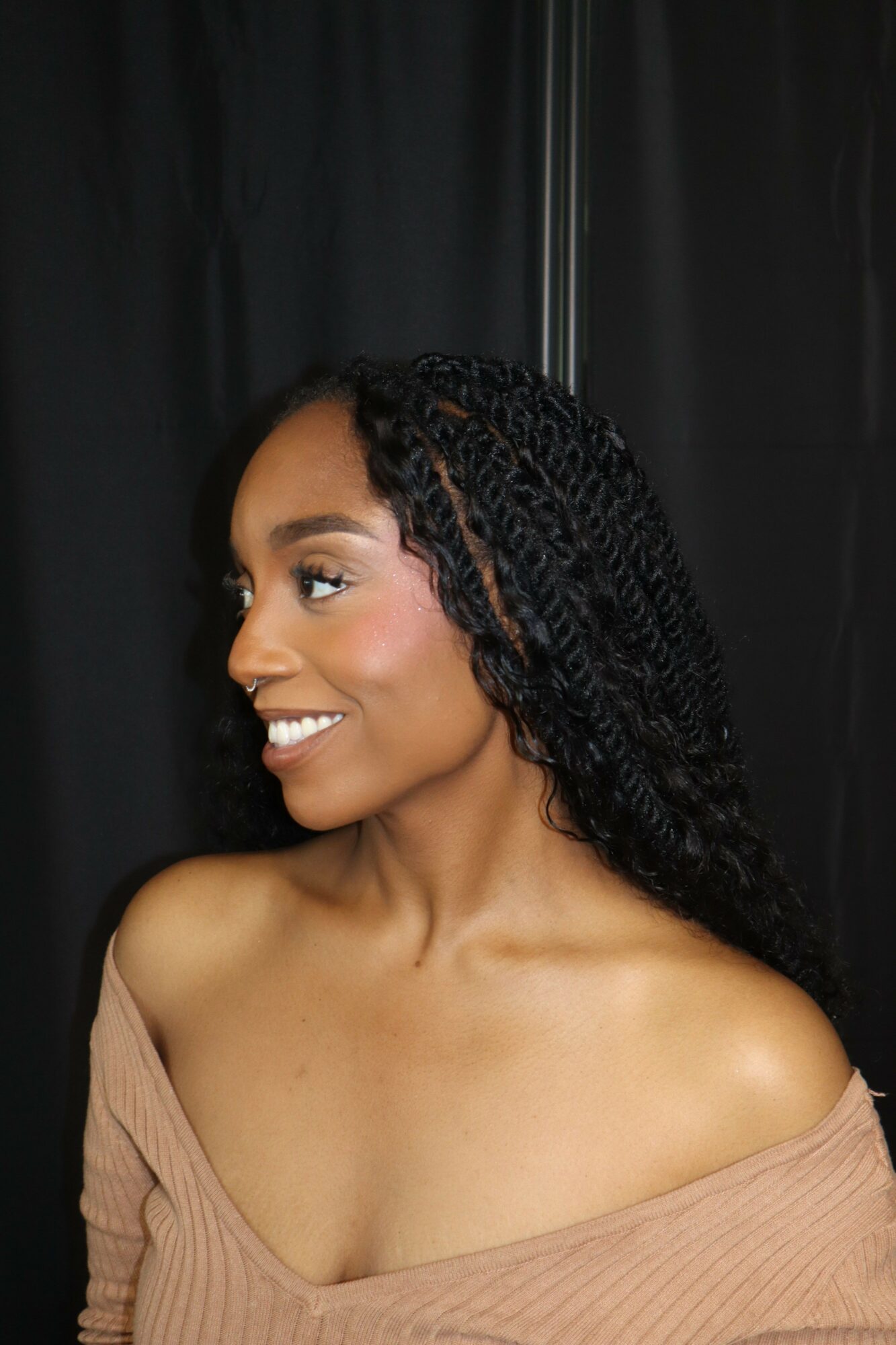 Woman with braided hair lying on a black surface, smiling, wearing a beige top, showing her shoulder and neck.