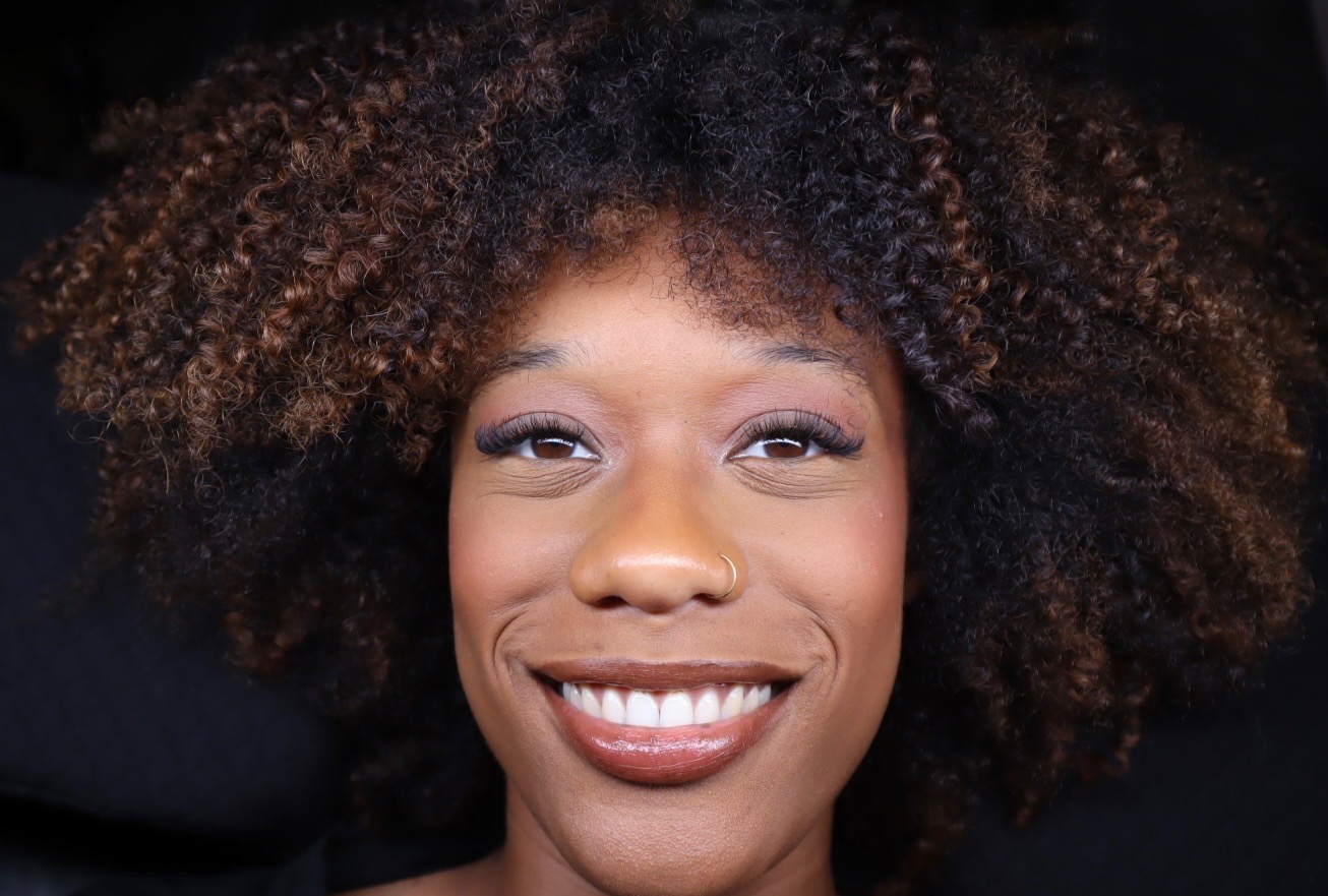 Close-up of a smiling woman with curly hair and makeup, looking directly at the camera.