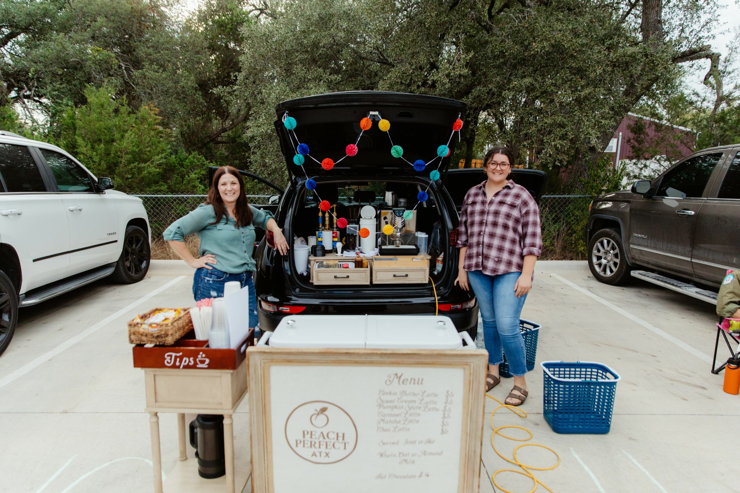 Two women stand next to a food truck with an open hatch, surrounded by parked cars and trees, in a parking lot.