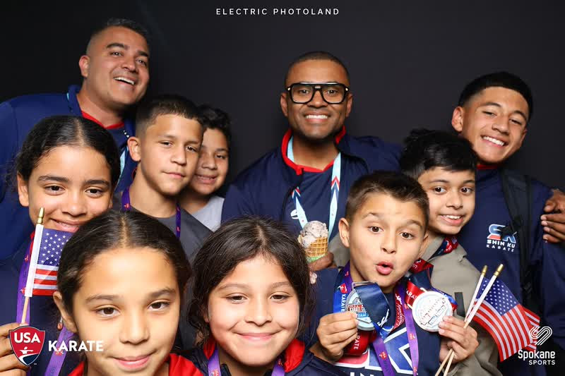 Group of smiling children and an adult holding medals and flags, standing together indoors against a dark background.