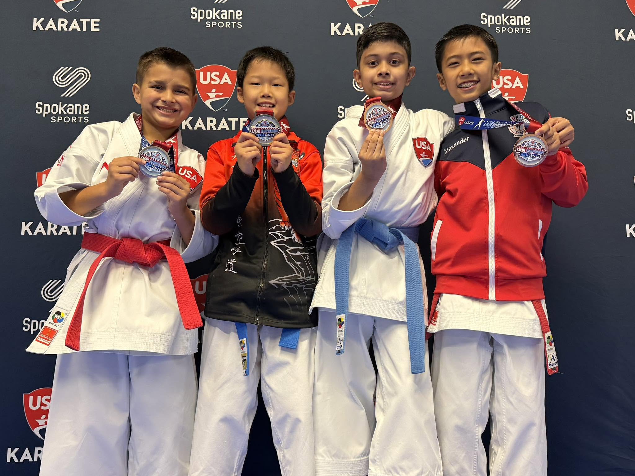 Four children in karate uniforms holding medals, standing in front of a backdrop with karate and USA logos.