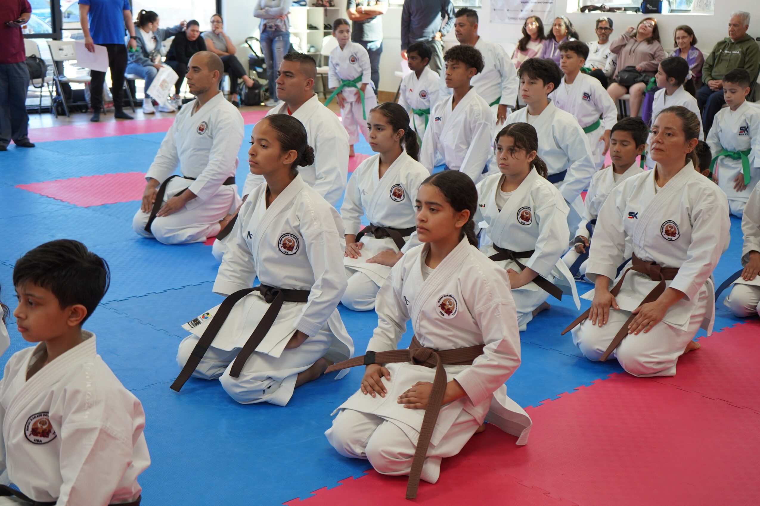 Group of children in martial arts uniforms sitting on mats during training or event, with spectators in background.