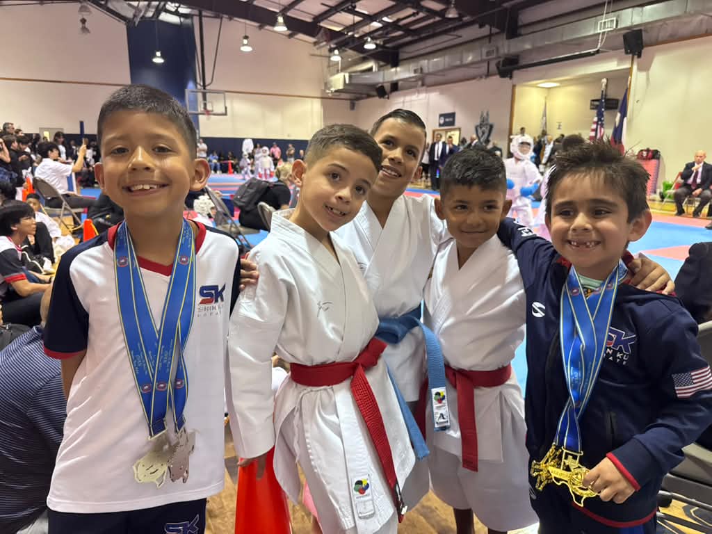 Five boys in martial arts uniforms and casual clothes, smiling with medals, in a gymnasium with spectators and equipment in background.