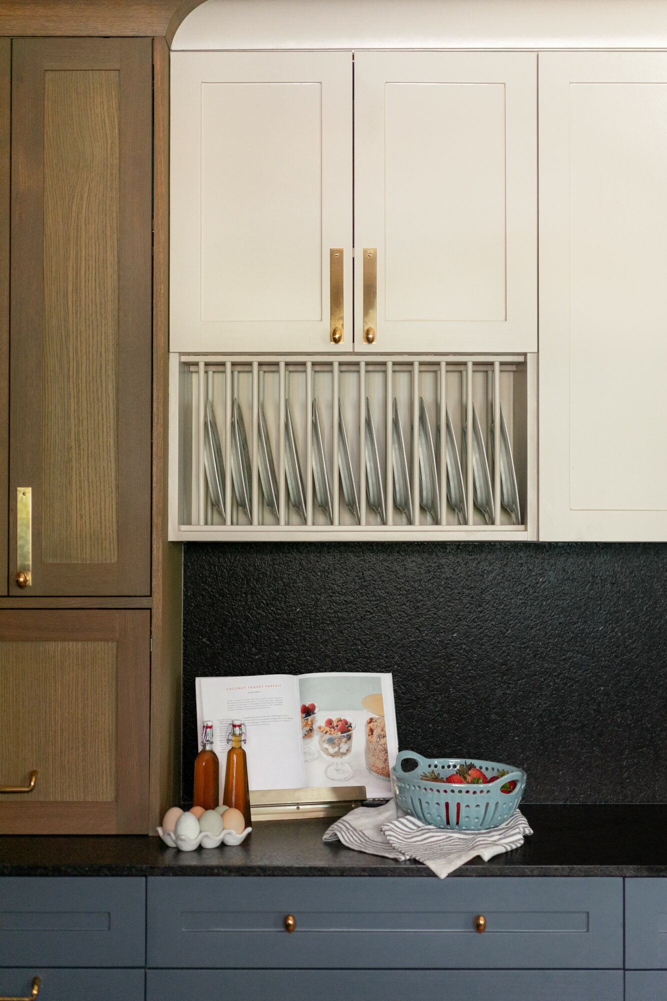 Kitchen cabinet with dish rack, countertop with bottles, a basket, and a book, against a black wall.