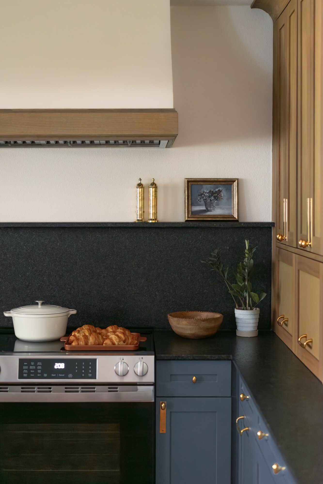 Kitchen countertop with a stove, pot, wooden bowl, potted plant, framed picture, and decorative bottles, with cabinets above and below.