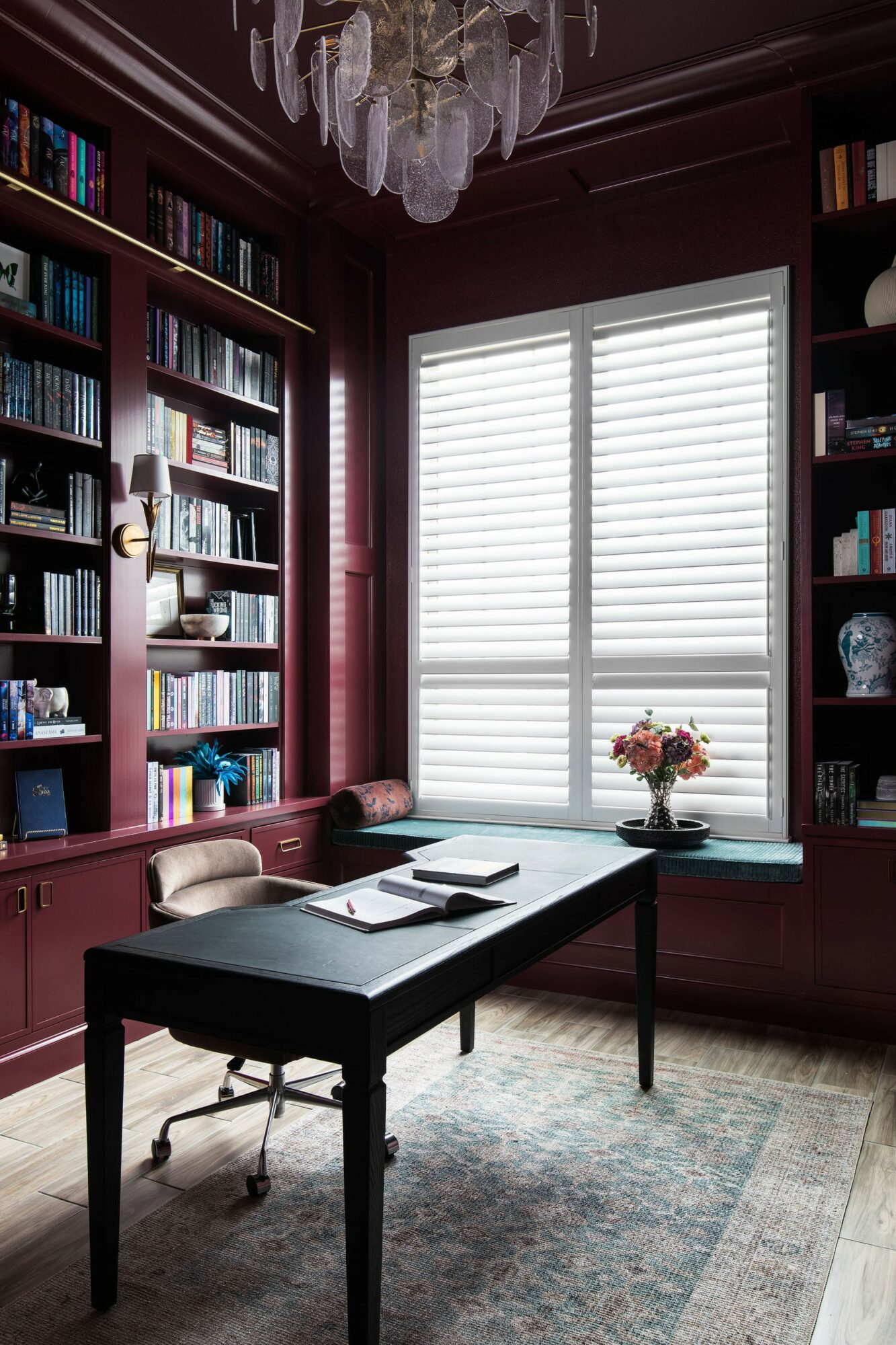 Office with dark wood bookshelves, a window with blinds, a desk with books and a flower vase, and a chandelier.