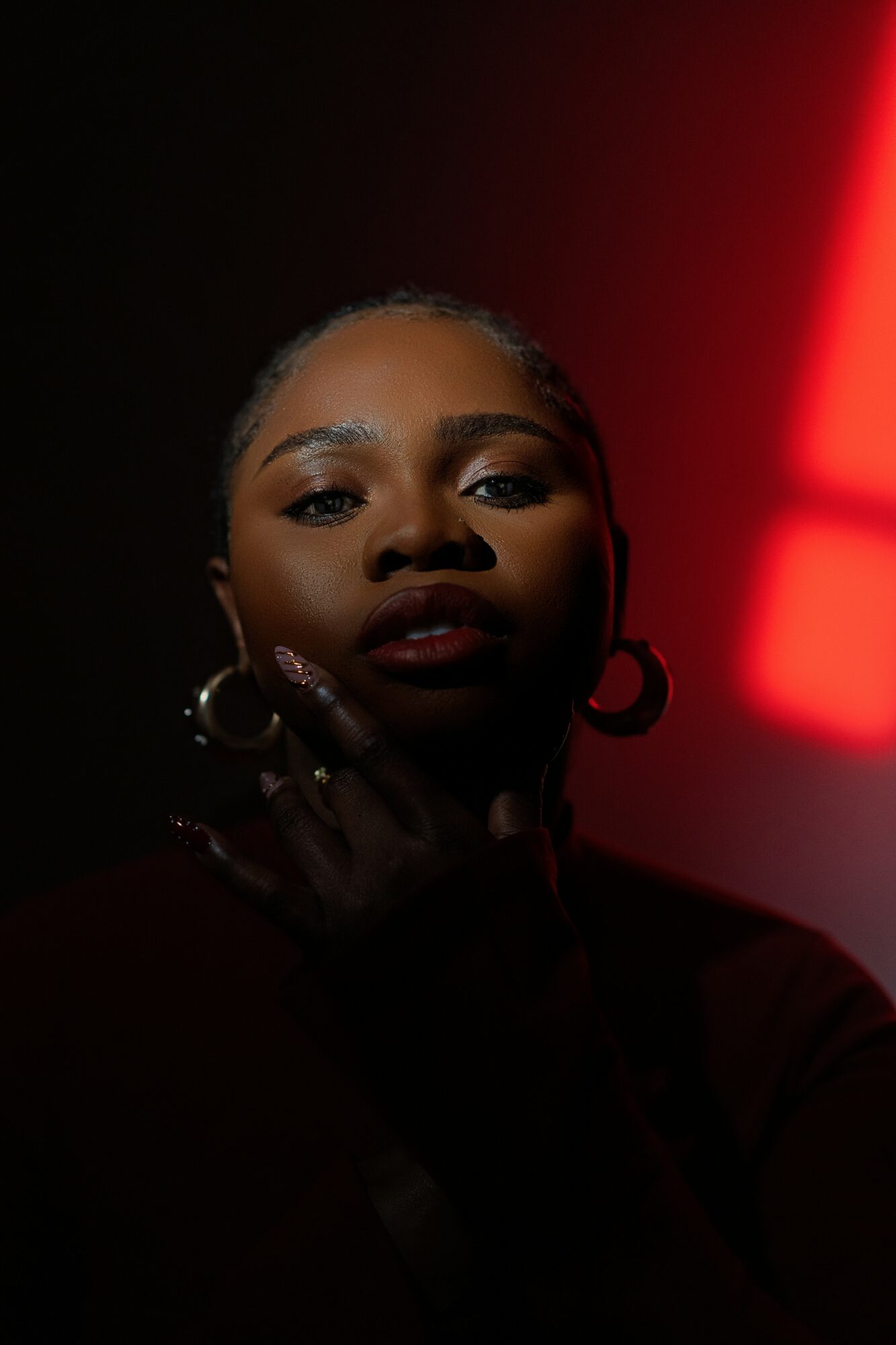 Portrait of a woman with hoop earrings, dark makeup, and a hand near her face, illuminated by red and black lighting.