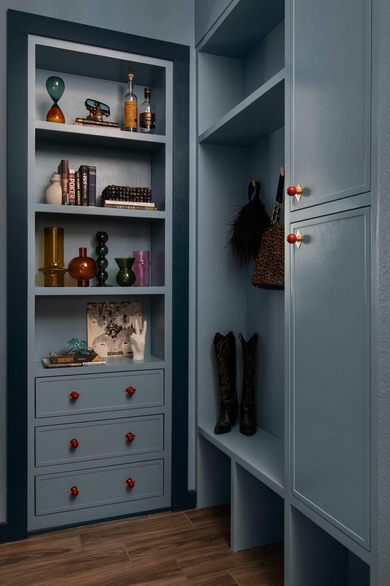 Blue built-in shelving unit with decorative items, books, and drawers, adjacent to a closet with hanging bag and boots, on wooden floor.
