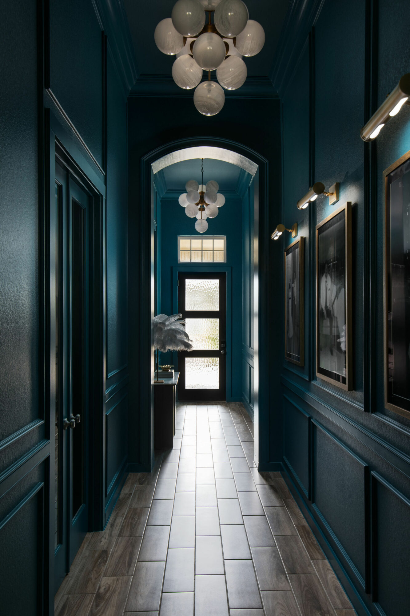 Narrow hallway with dark teal walls, framed artwork, and a chandelier hanging from the ceiling, leading to a door with windows.