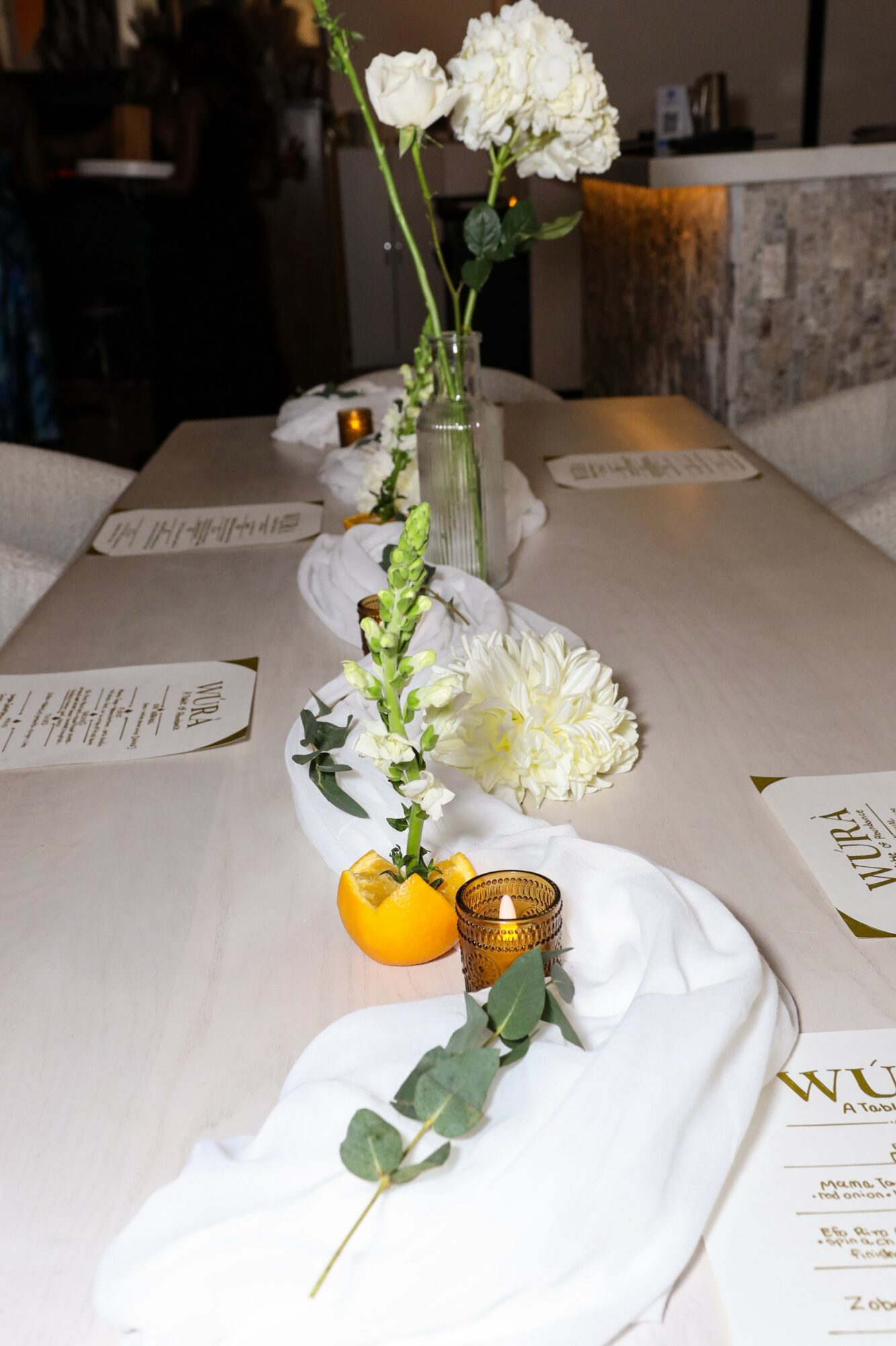 Table with white flowers in a glass vase, candles, and a white cloth runner, set for a meal.