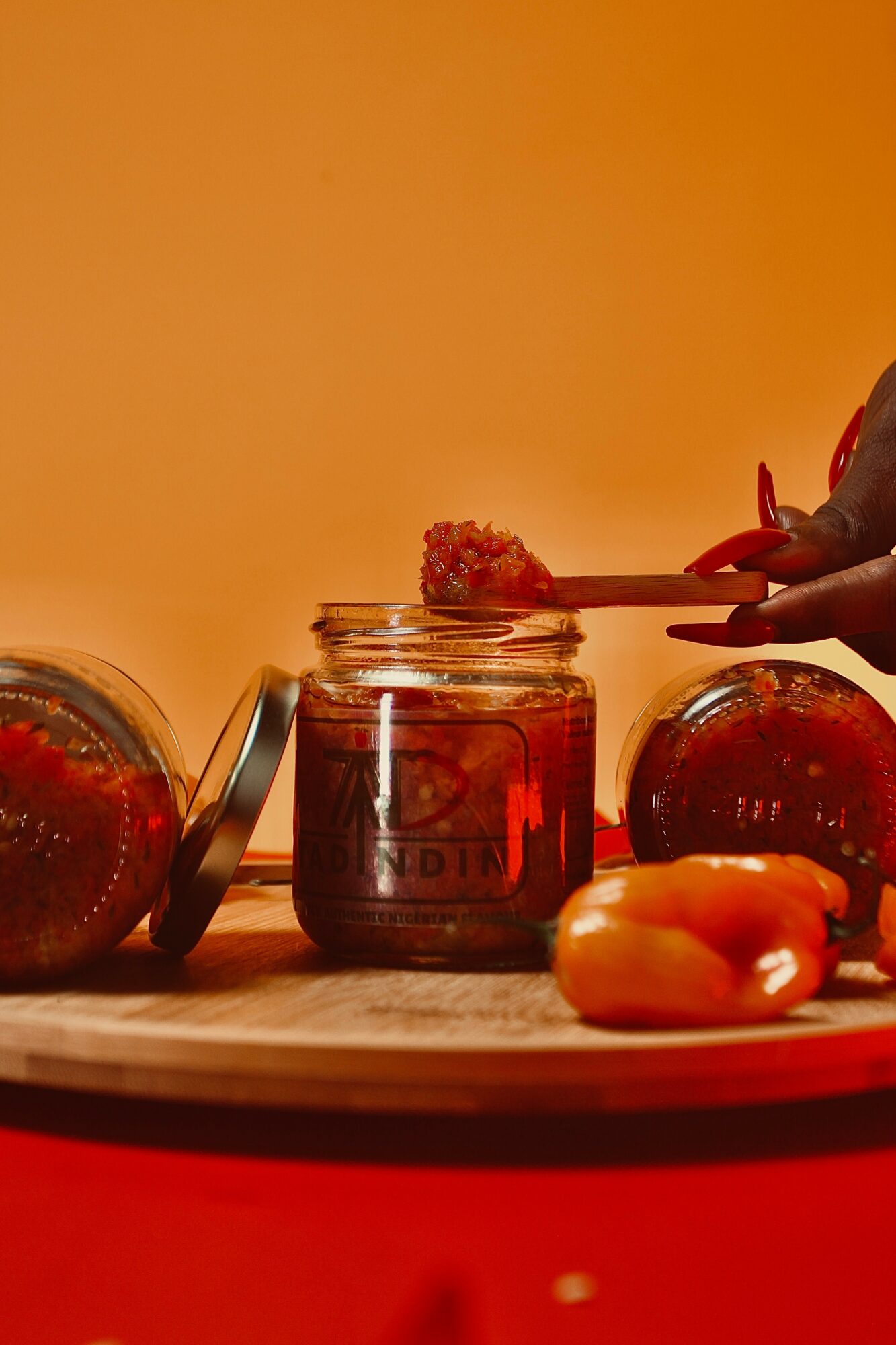 Three jars of jam, a spoon with jam, and some apricots on a wooden surface against an orange background.