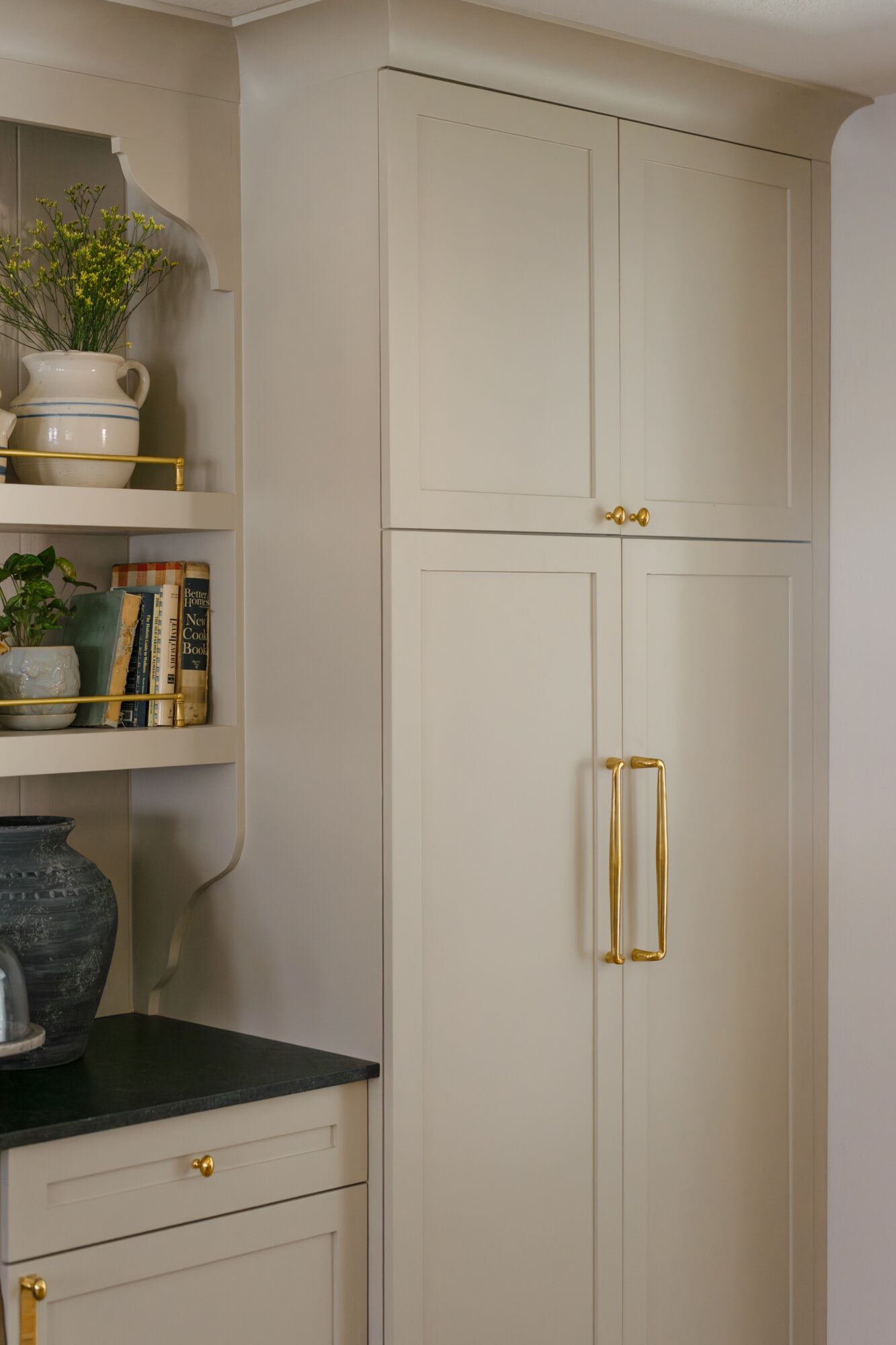 Tall white cabinet with gold handles in a room corner, next to a shelf with decorative items and books.