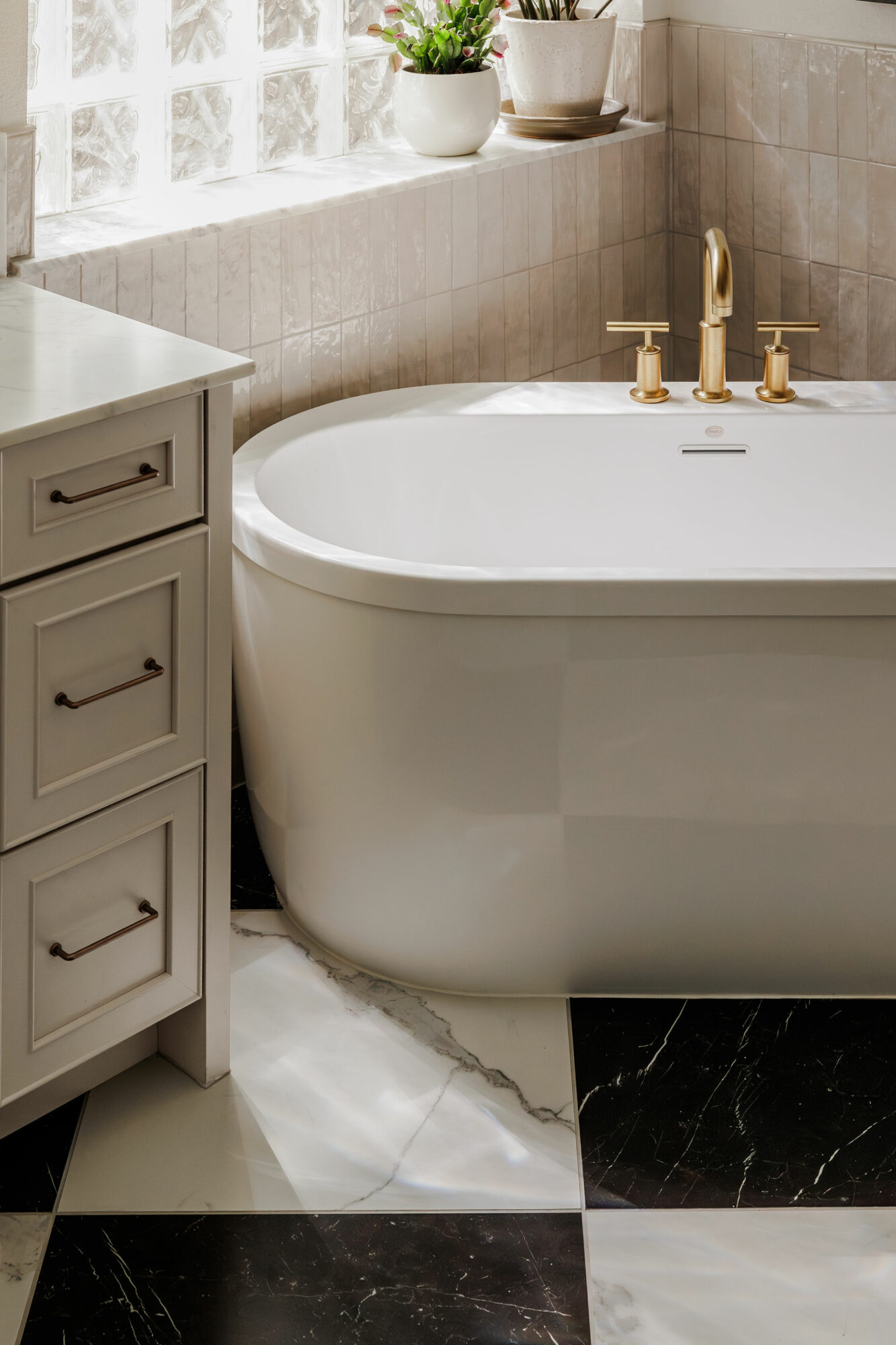 Bathtub with gold fixtures next to a gray cabinet, with potted plants on a windowsill, tiled wall, and checkered floor.