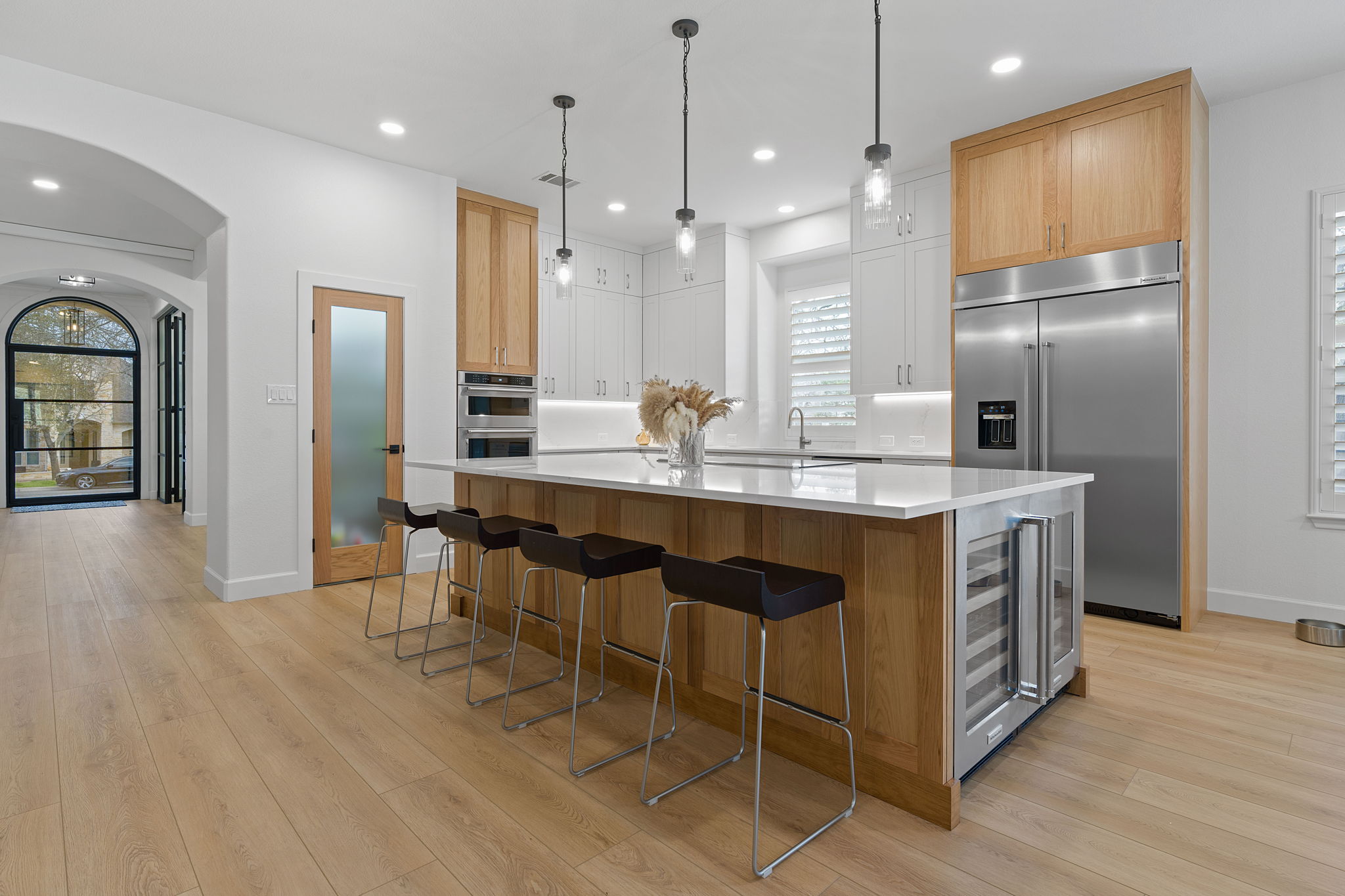 Modern kitchen with island, pendant lights, wooden cabinets, stainless steel refrigerator, and light hardwood floors.