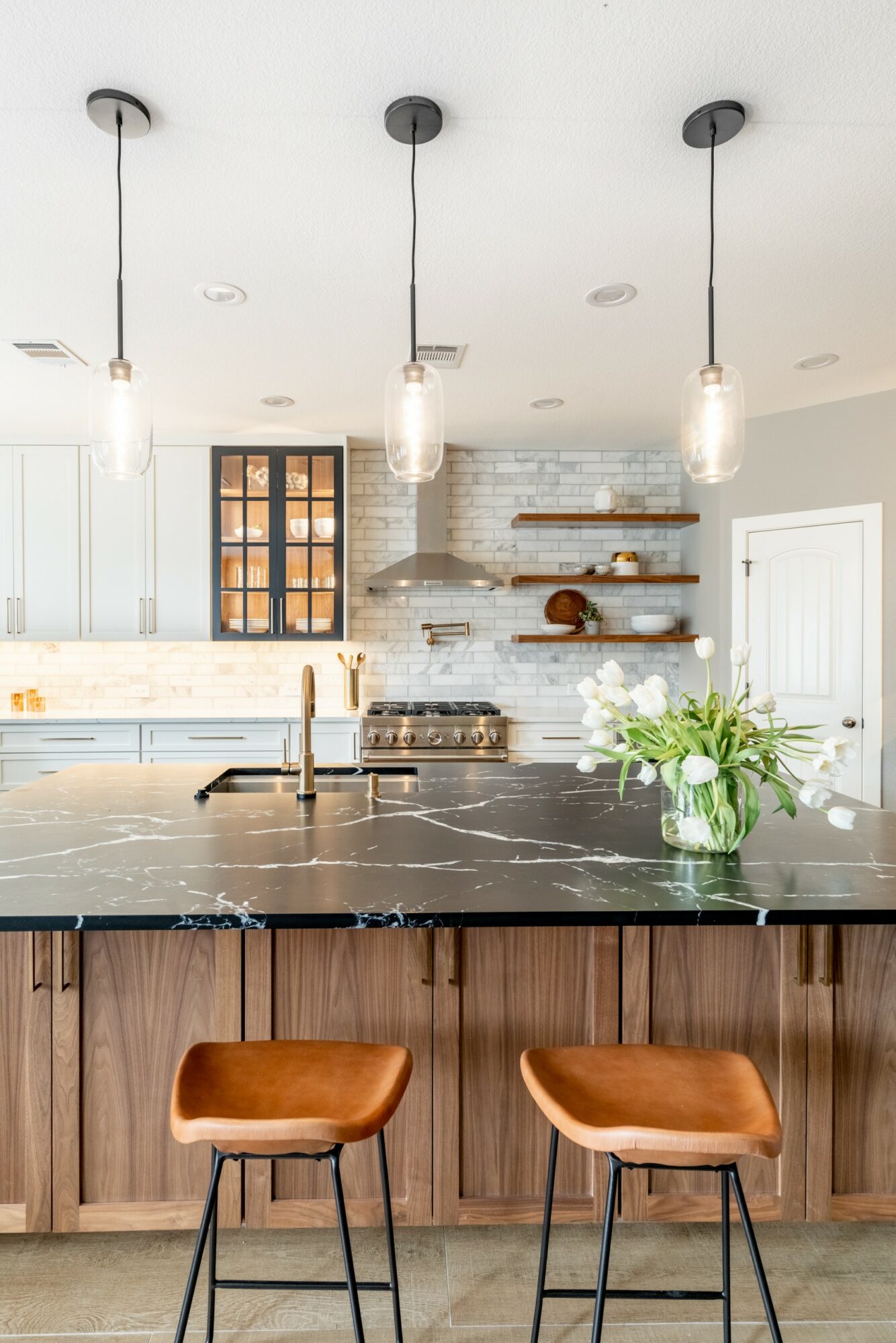 Modern kitchen with pendant lights, open shelves, and a black marble island with a vase of flowers, wooden bar stools, and a brick wall.