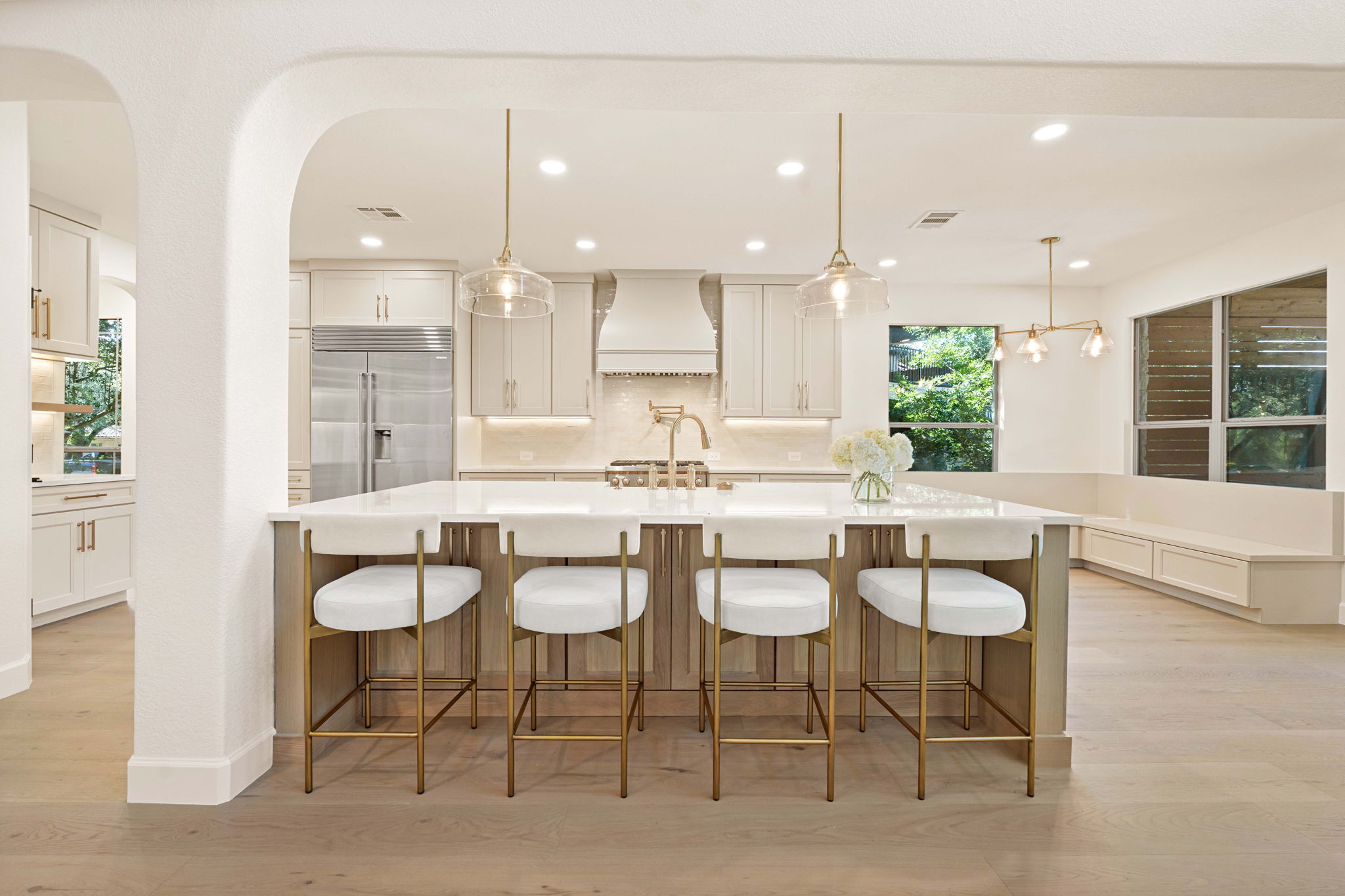 Modern kitchen with white cabinets, island with four white stools, pendant lights, and large windows, bright and spacious.