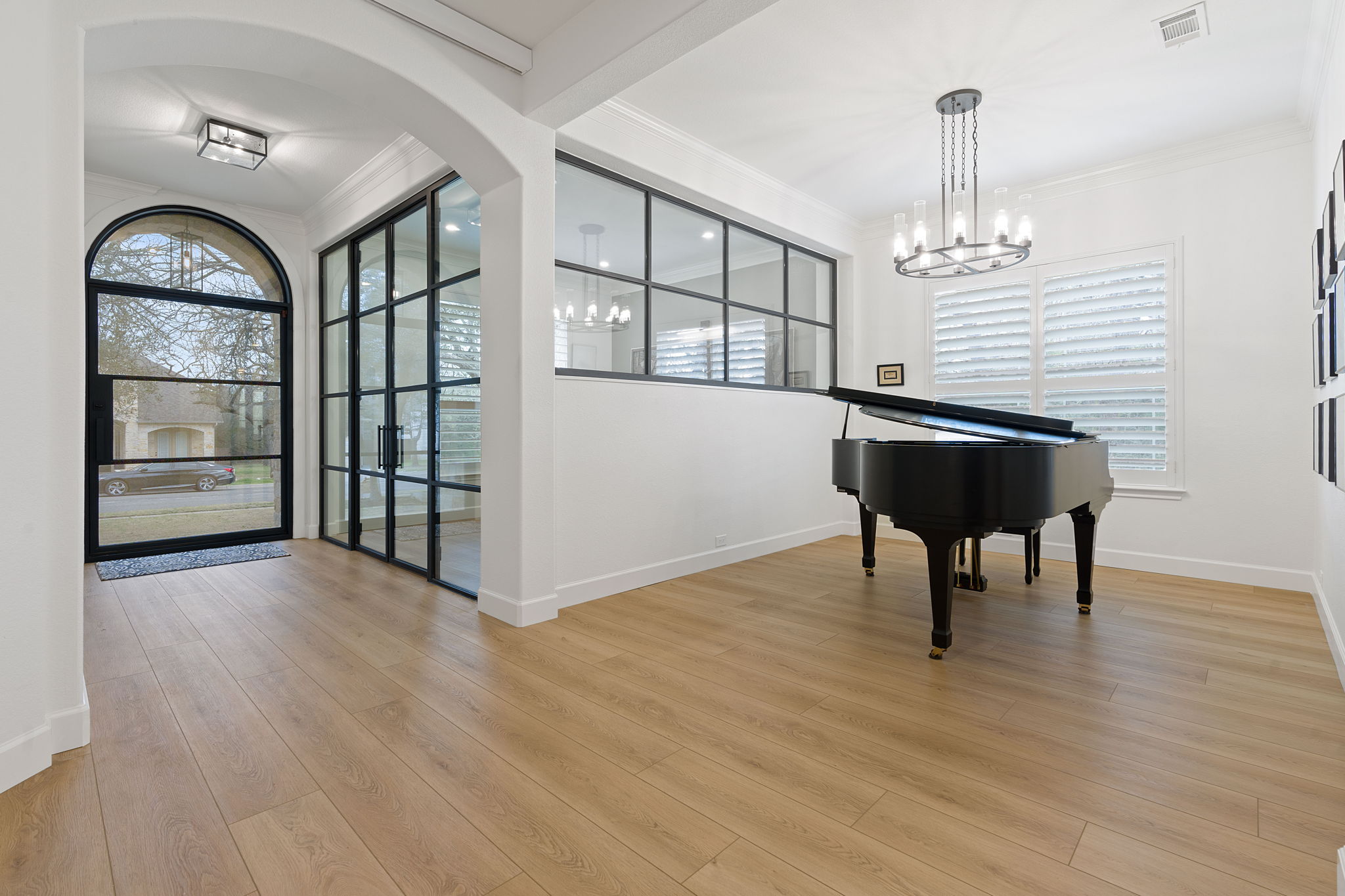 Room with a grand piano, large windows, and a chandelier, featuring light wood flooring and white walls.
