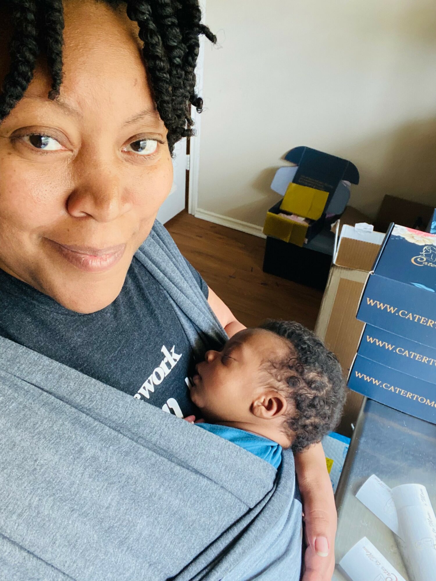 Woman holding a sleeping baby in a room with a chair and boxes in the background.
