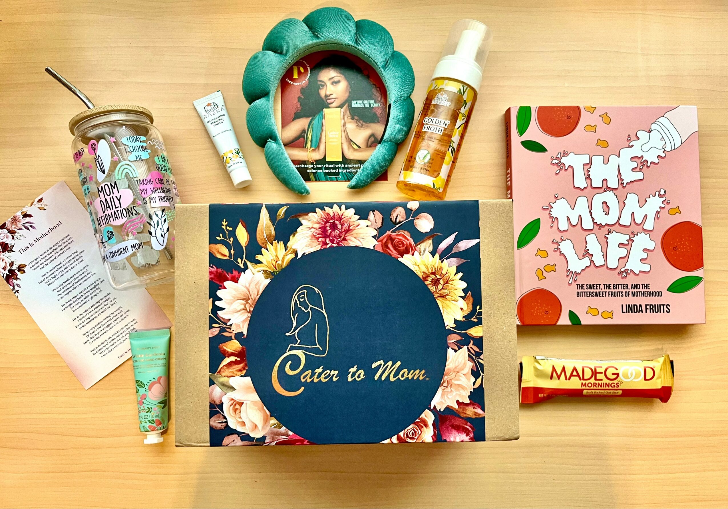 Flat lay of a gift box, skincare products, a book titled 'The Mom Life,' and a photo frame with a woman, on a wooden surface.