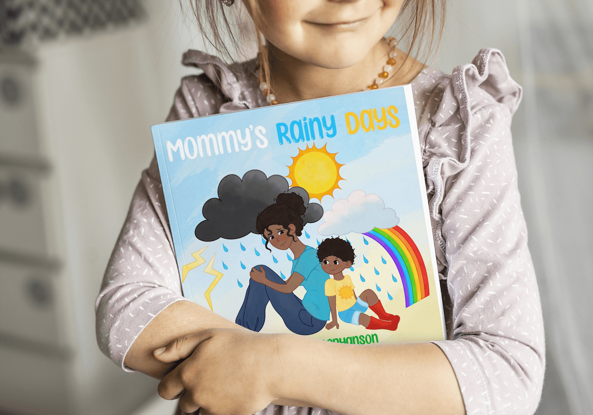 Child holding a book titled 'Mommy's Rainy Days' with illustrations of a girl, rainbow, and weather.