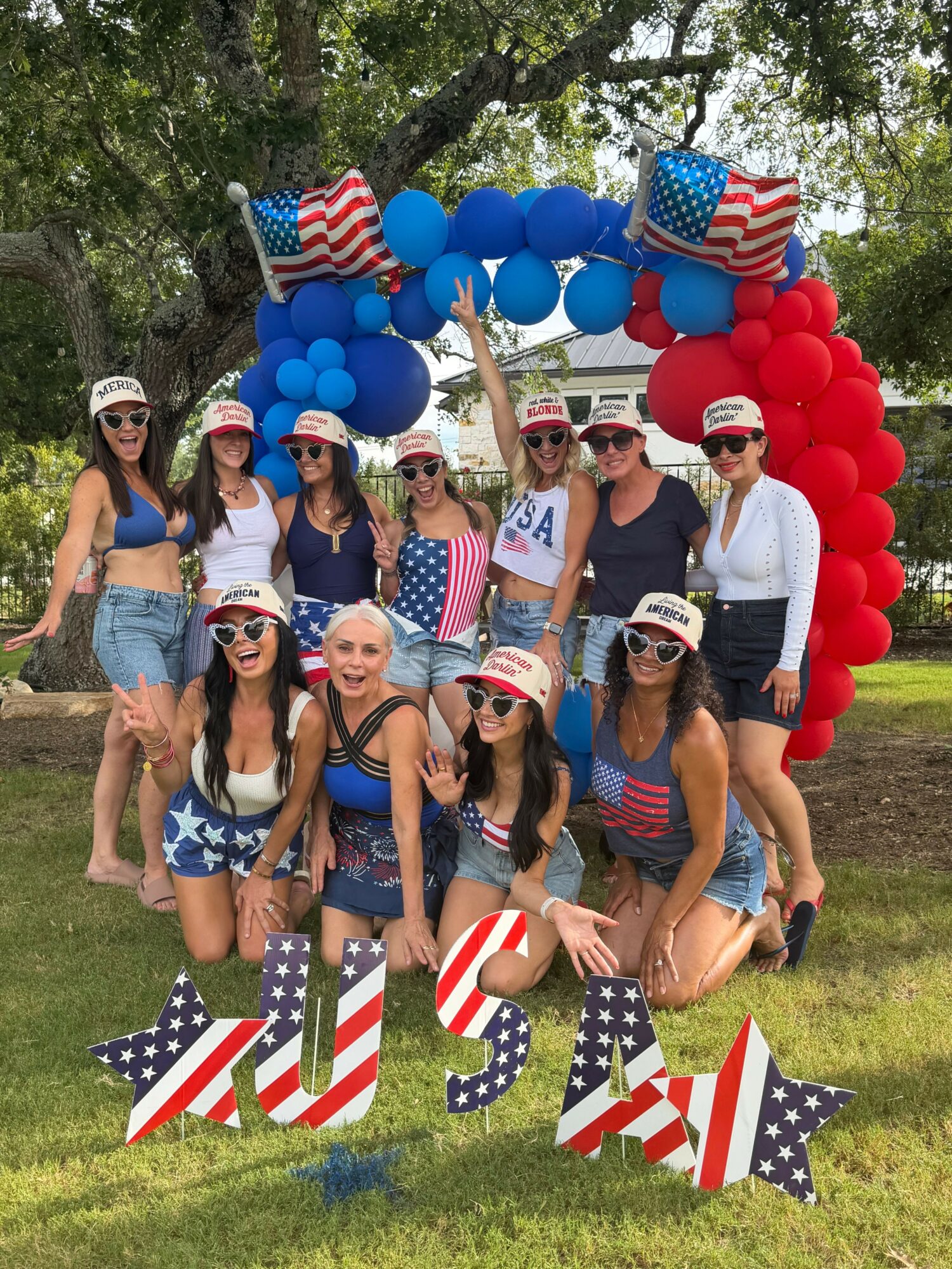 Group of people celebrating outdoors with patriotic decorations, balloons, and American flags, some wearing patriotic clothing, on grass with trees.