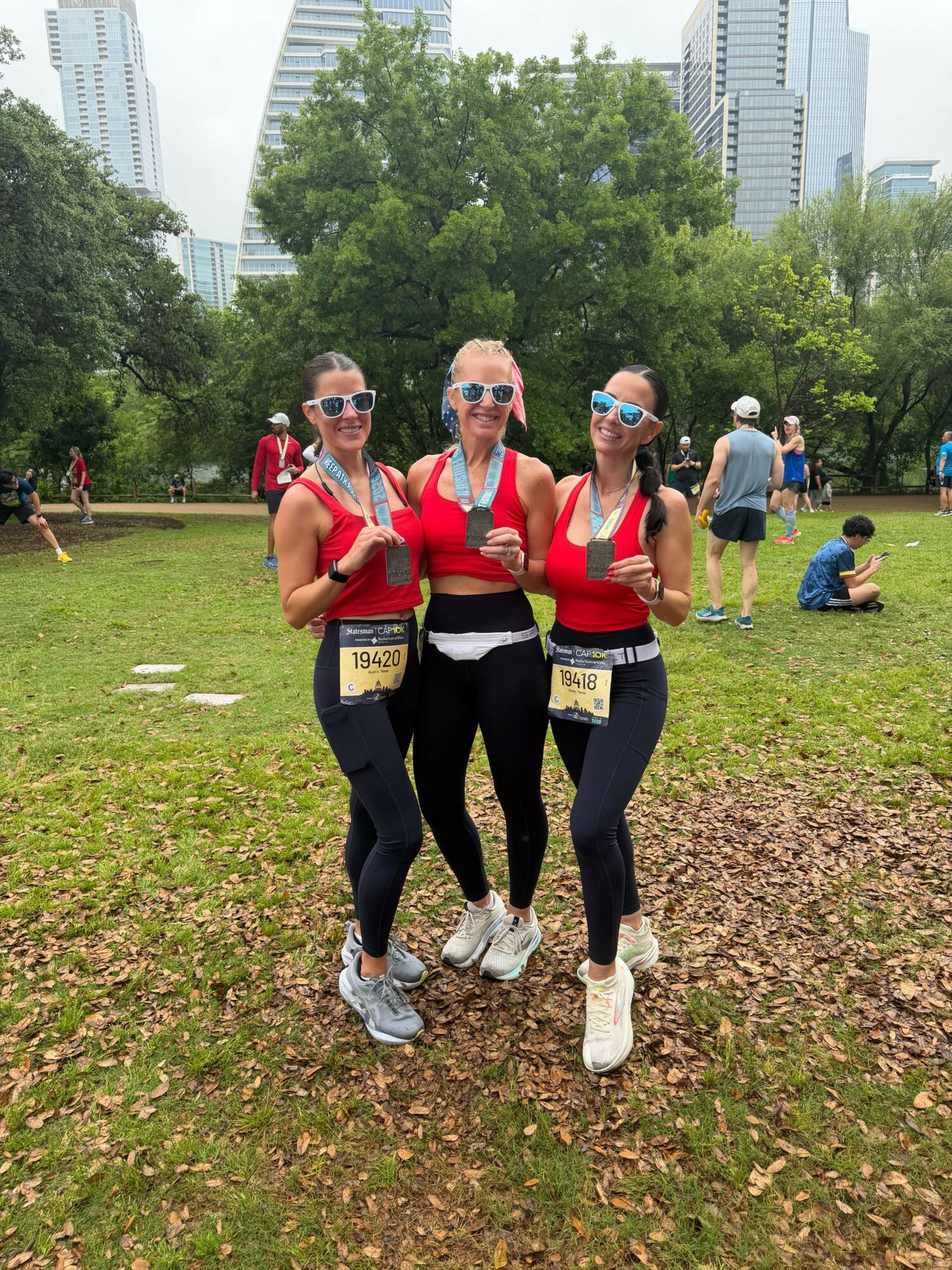 Three women in athletic clothing running outdoors on a grassy area with trees and buildings in the background.