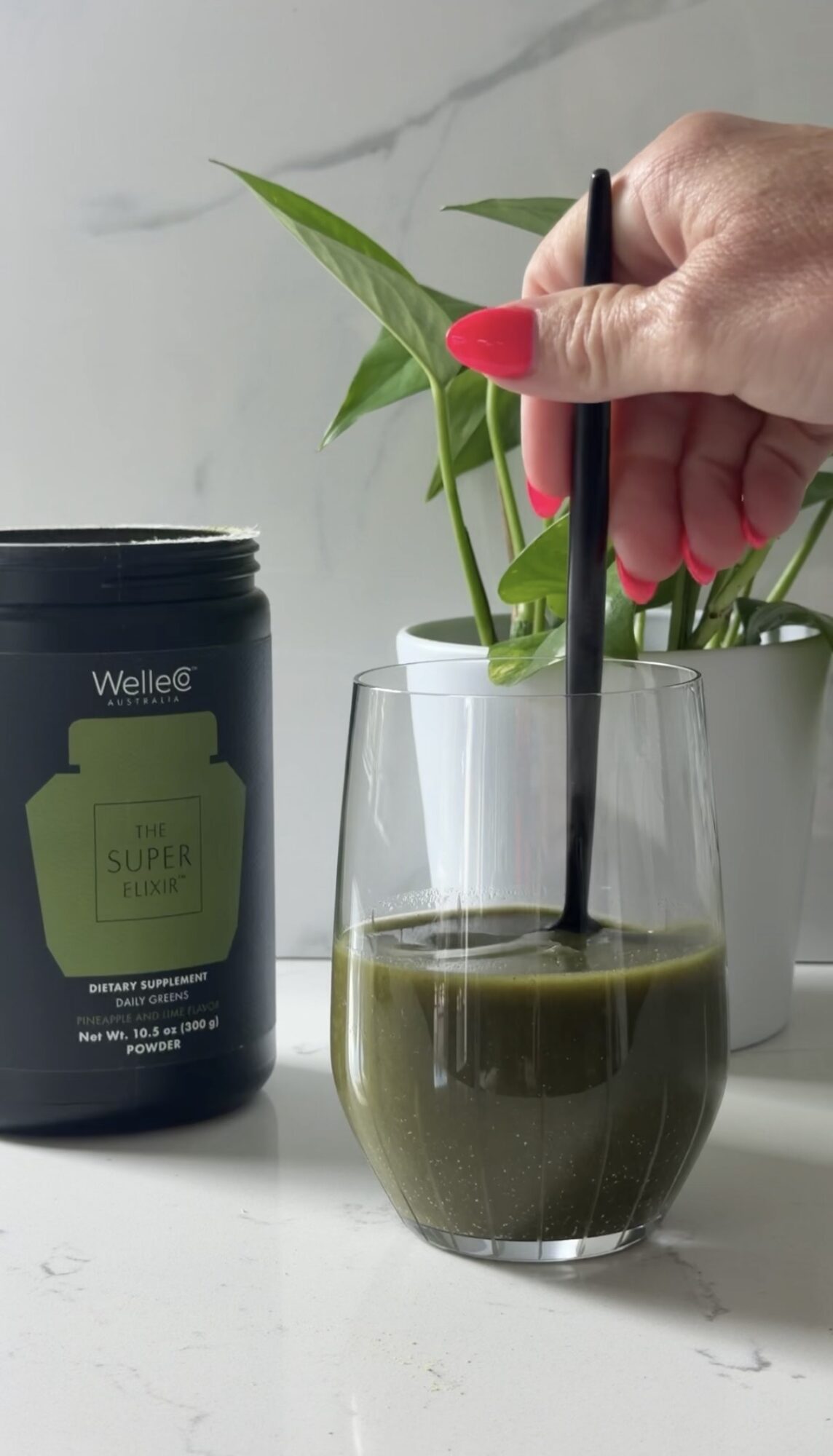 Hand with red nail polish holding a black straw in a glass of dark green liquid, with a plant and container nearby.