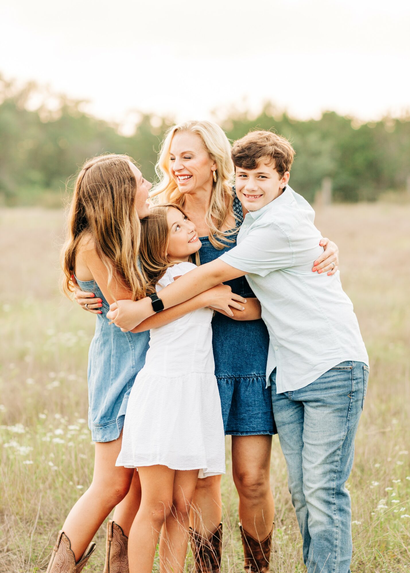 A woman and three children hugging outdoors in a grassy field, smiling, with trees in the background.