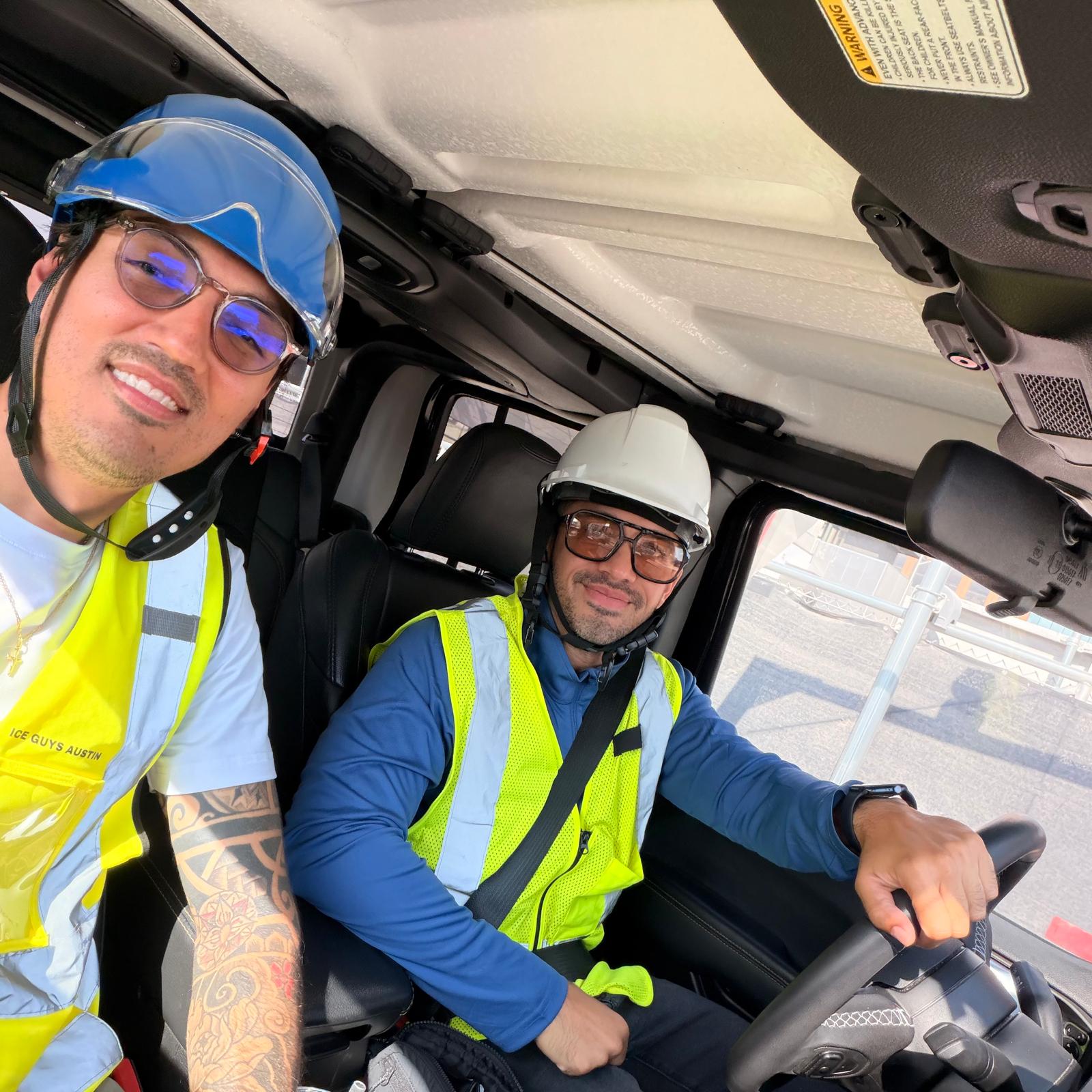 Two men in safety vests and helmets inside a vehicle, one driving and one taking a selfie.