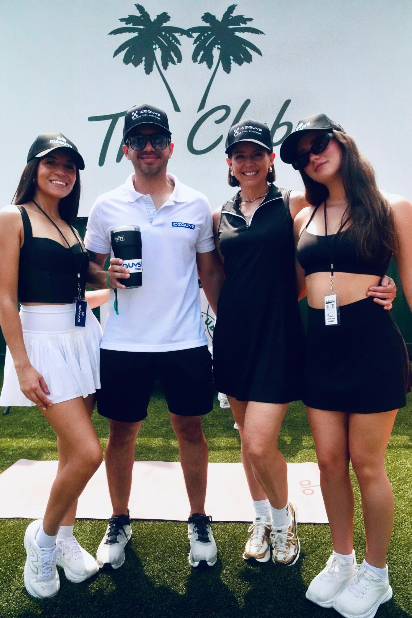 Four people standing outdoors in front of a sign with palm trees, smiling, wearing casual summer clothing and sunglasses.