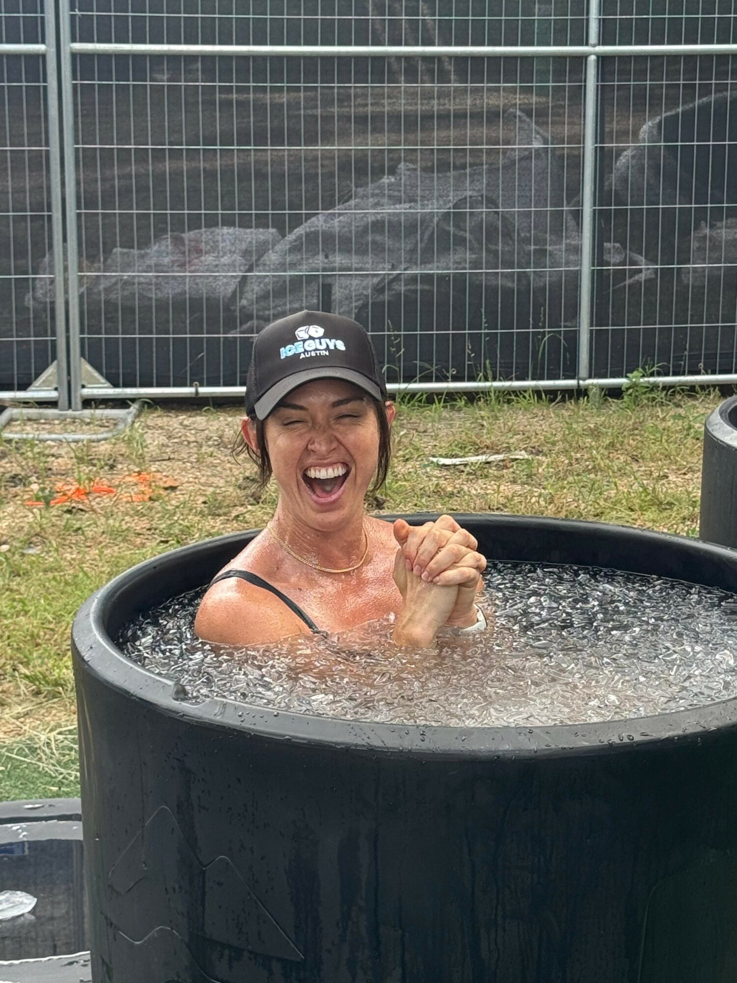 Woman smiling and laughing in a black tub outdoors, with a fence and rocks in the background.
