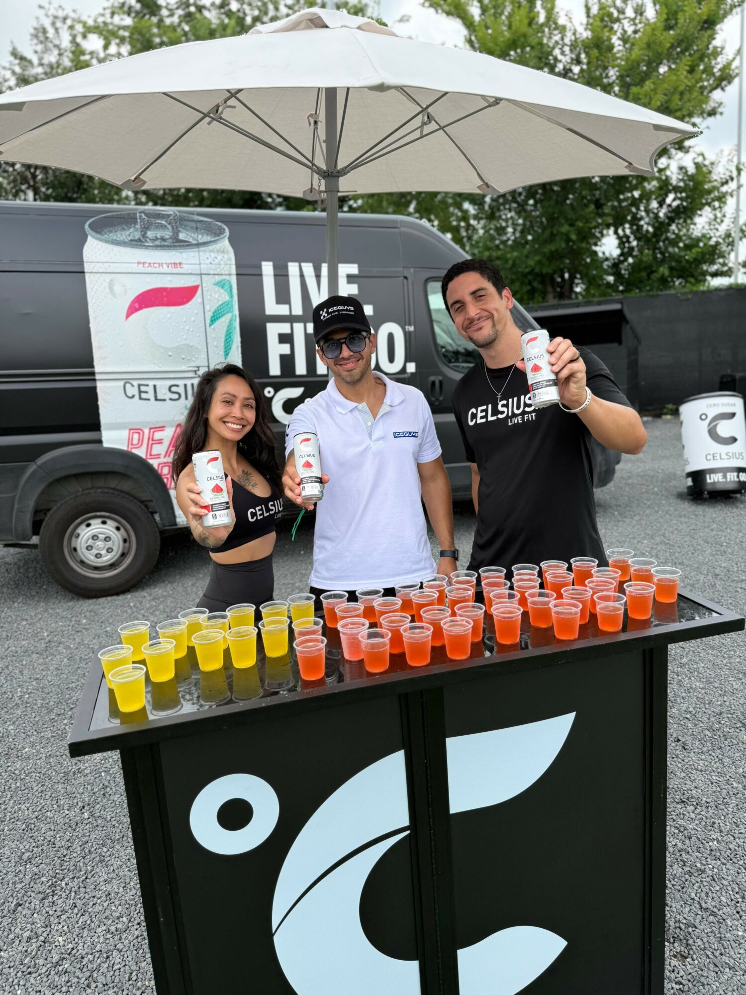 Three people standing behind a table with colorful drinks, under a large white umbrella, outdoors with trees and a vehicle in background.
