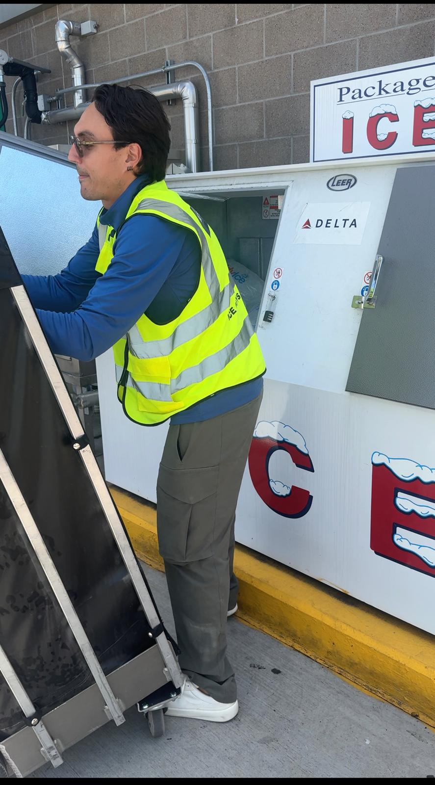 Person in a yellow safety vest using a machine outside near ice vending machines, brick wall background.