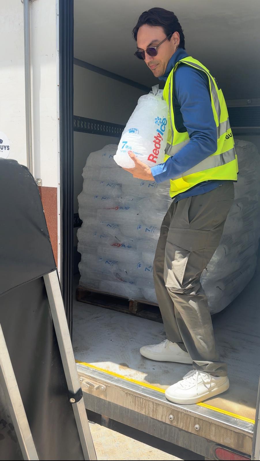 Man wearing sunglasses, a yellow safety vest, and gray pants, holding a bag of supplies near stacked items.