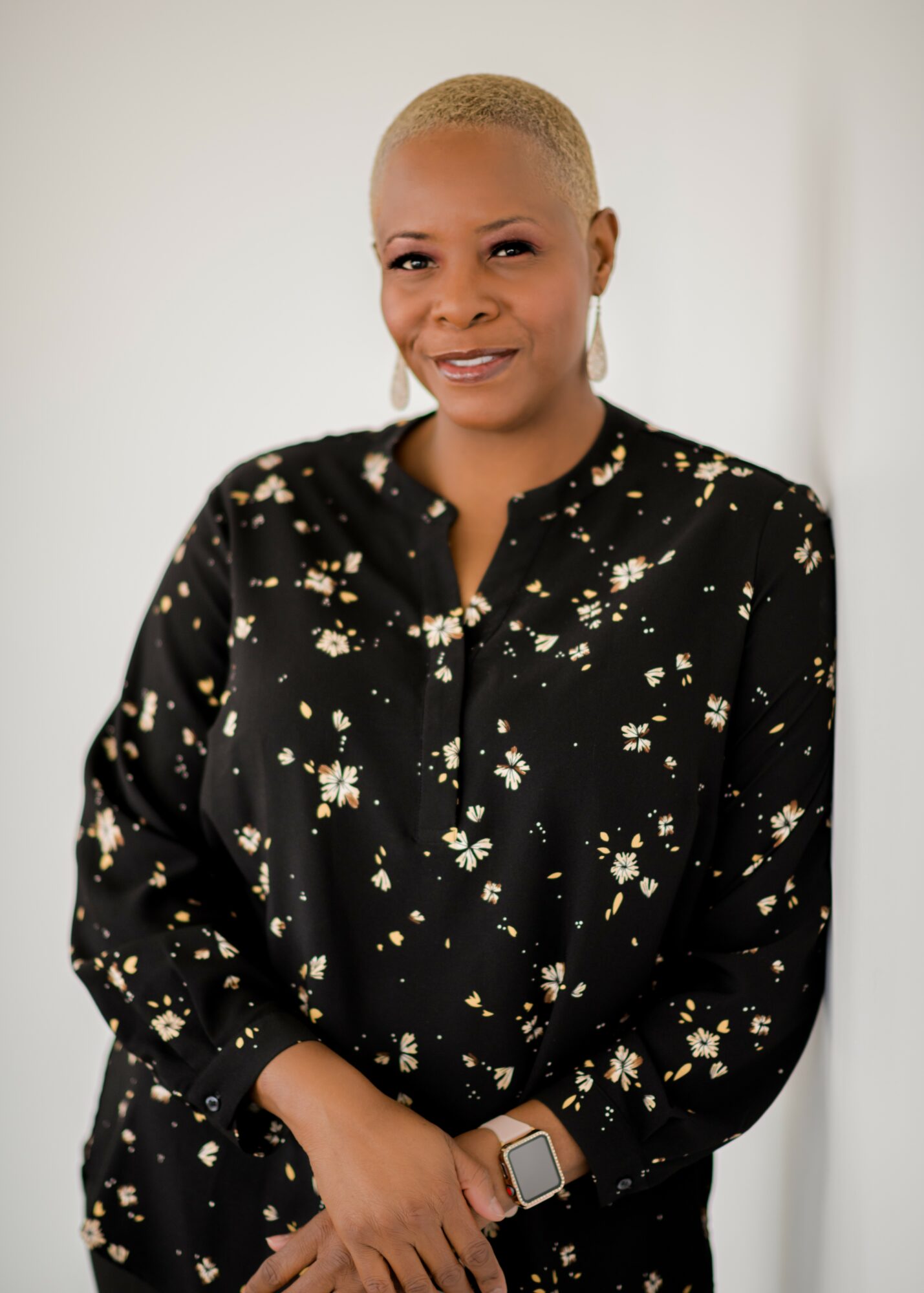 Woman with short blonde hair wearing a black floral blouse, smiling, standing against a plain background.