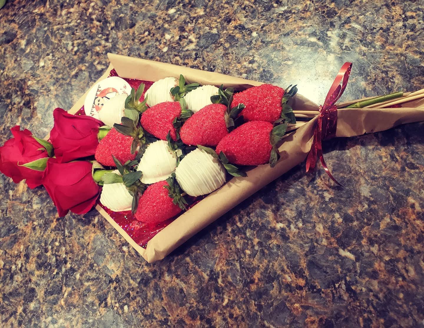Bouquet of red roses and strawberries wrapped in paper with a red ribbon on a dark granite surface.