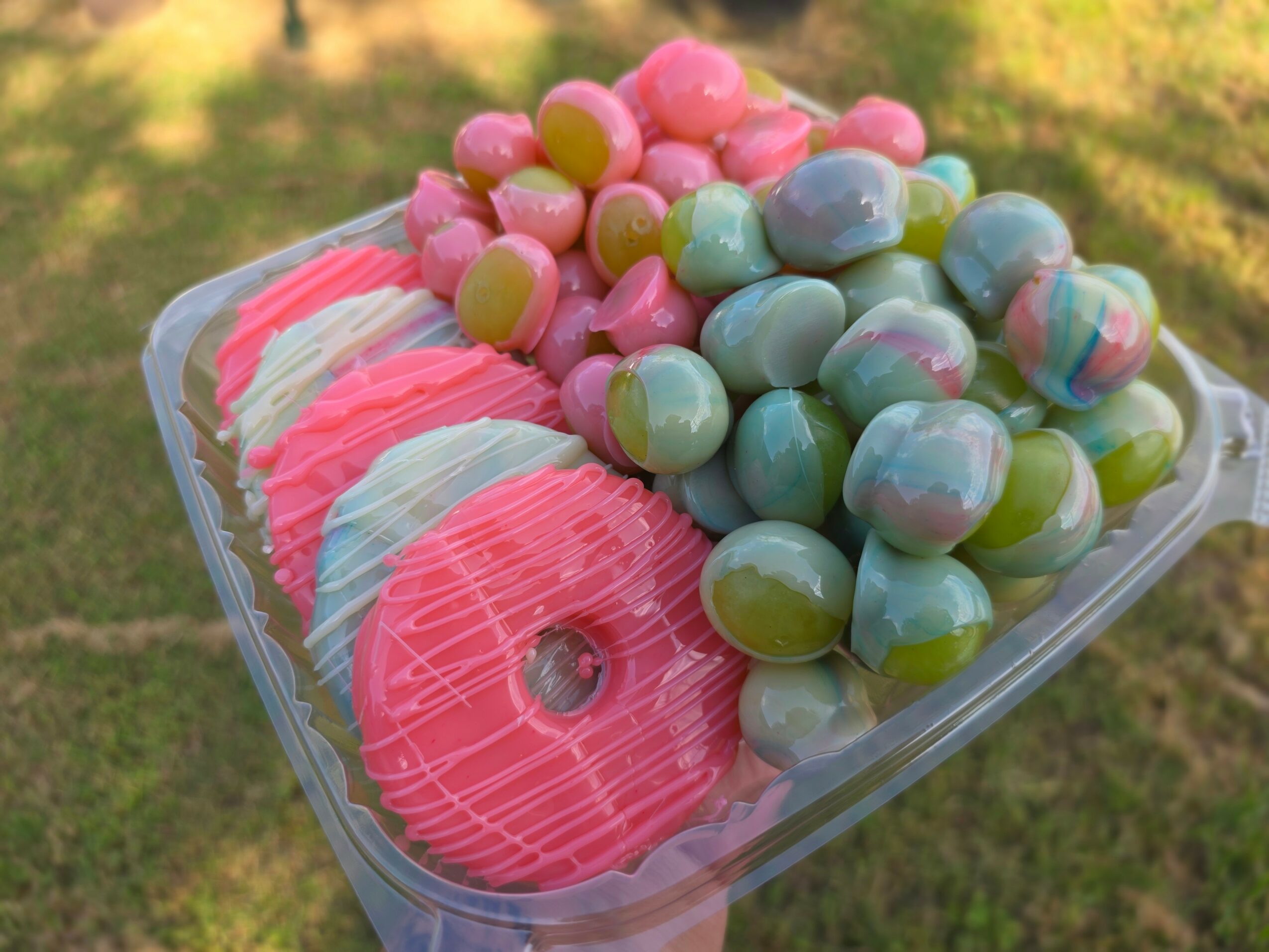Clear container with pink, green, and marbled candies, including donuts and round candies, outdoors on grass