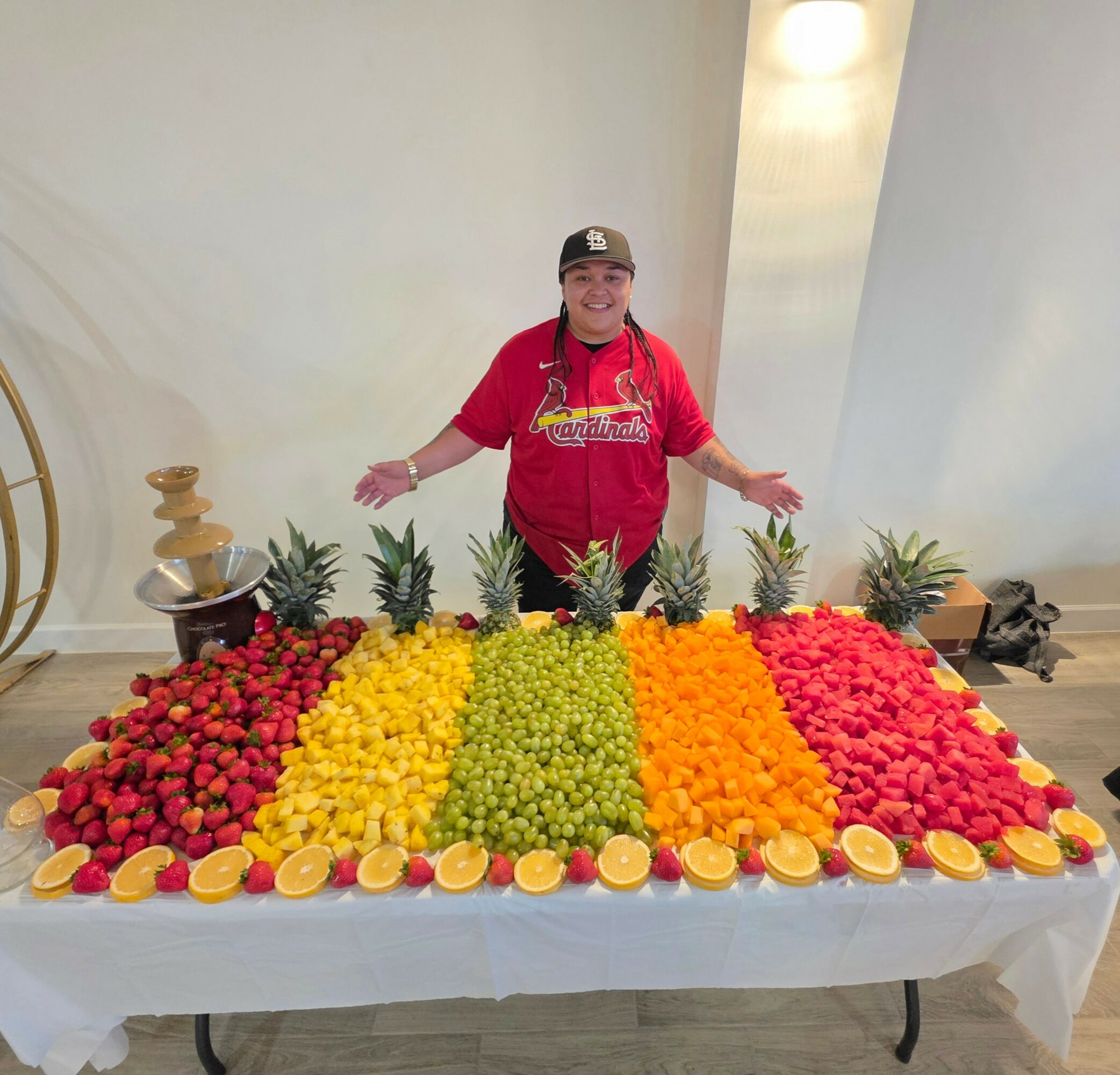 Person standing behind a table with colorful fruit arranged in rows, with pineapples and lemon slices, in a bright room.